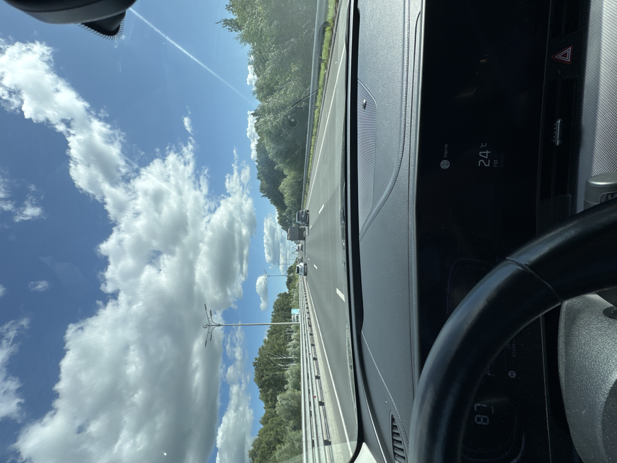 The image, taken from the driver's perspective inside a vehicle, shows a highway scene under a partly cloudy blue sky. The road has multiple lanes and is bordered by guardrails and lush green trees on both sides. Several vehicles are visible on the highway, including cars and a truck. In the foreground, the car's dashboard and steering wheel are visible. The digital display shows a speed of 87 km/h and a temperature of 24°C, indicating FM radio is playing. The location is identified as Kazan, Russia.