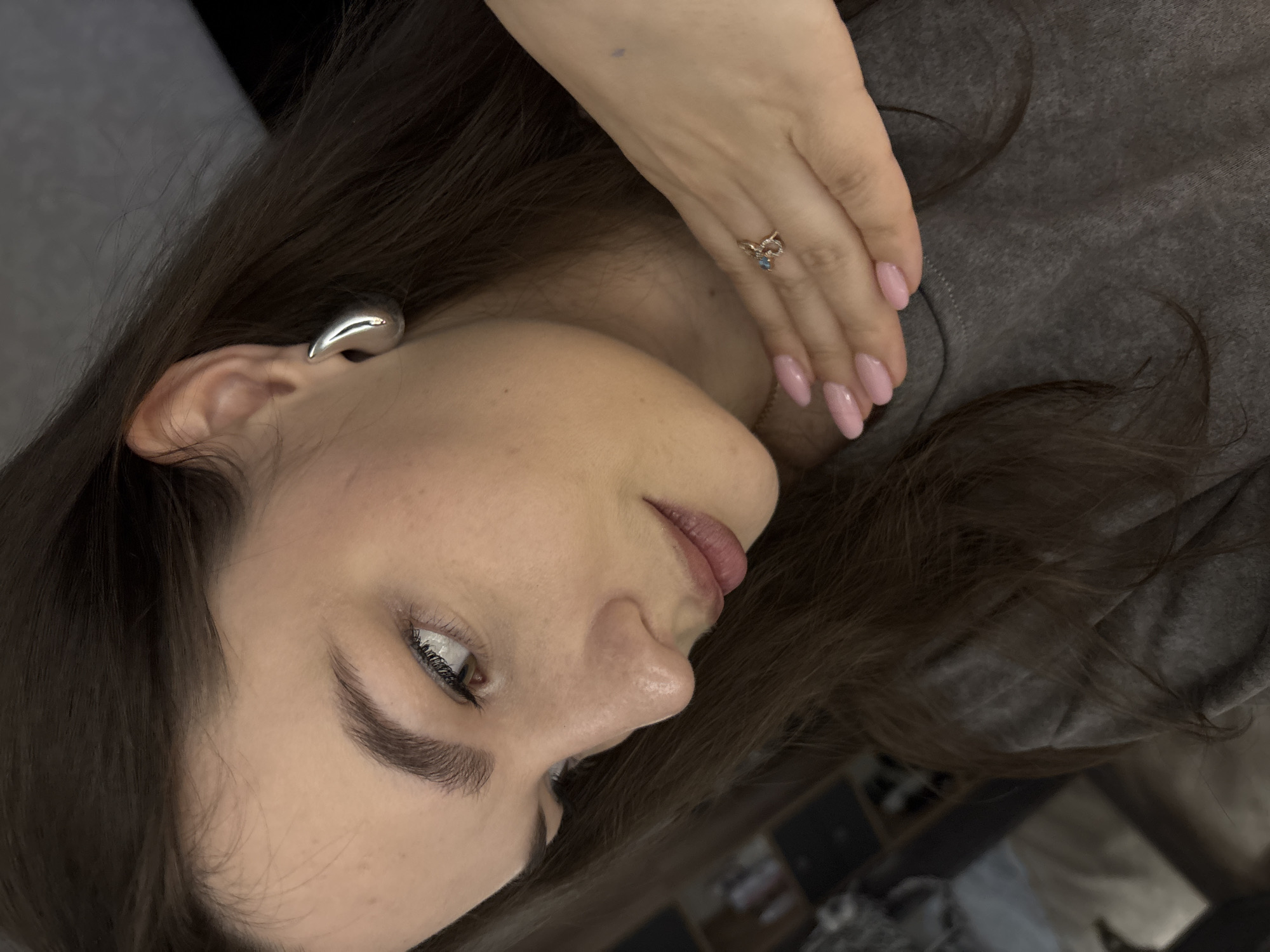 A close-up, slightly rotated image depicts a woman with light skin, dark brown hair, and a neutral expression. Her right ear is visible, adorned with a large, silver-colored ear cuff. Her left hand, with light pink manicured nails, rests near her face, featuring a ring with a blue stone. The woman wears a gray top. The background is blurred, suggesting an indoor setting. The location in Chaikovsky, Russia, cannot be determined from the image alone.