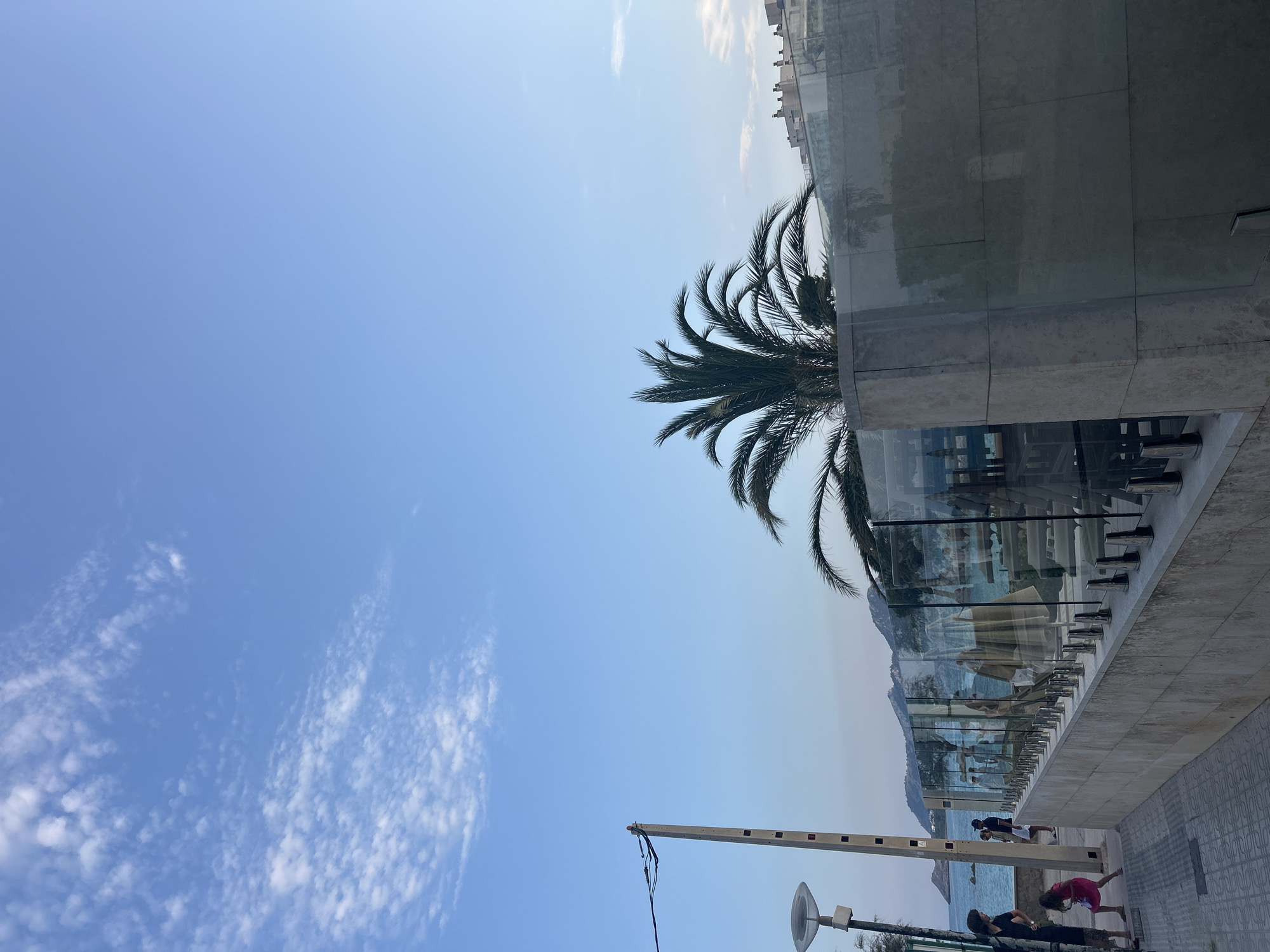 The image captures a coastal scene in Capdepera, Spain, likely near the sea. The sky is a clear, pale blue with scattered wispy white clouds. In the foreground, a paved walkway runs along a textured stone wall topped with a glass railing system. Several people are visible on the walkway; a woman and a child in a pink dress are in the lower left, with two other individuals further down the path. Behind the glass railing, a tropical palm tree with fronds extends into the frame, and beyond that, the calm blue sea and distant, hazy hills are visible. A building with multiple windows and a patterned roofline is situated on the hillside in the background. A light pole with electrical wires is also present on the left side of the walkway. The lighting suggests it is late afternoon or early evening.