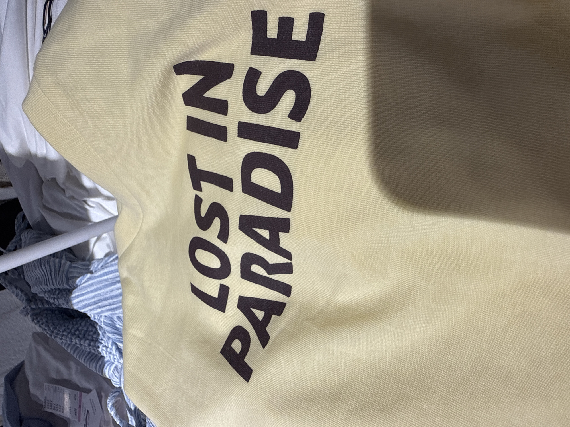 This media file shows a close-up, vertically oriented view of a light yellow t-shirt with dark brown text printed on it. The text reads "LOST IN PARADISE" in a bold, sans-serif font, with the words stacked vertically. The fabric of the t-shirt appears to be a textured knit. The lighting suggests an indoor setting, possibly a room or closet, with a shadow cast across the right side of the t-shirt. In the background, out of focus, there are other garments and what appears to be a white metal bar, potentially part of a clothing rack. There are no people visible in the frame. The focus is entirely on the t-shirt and its graphic print.