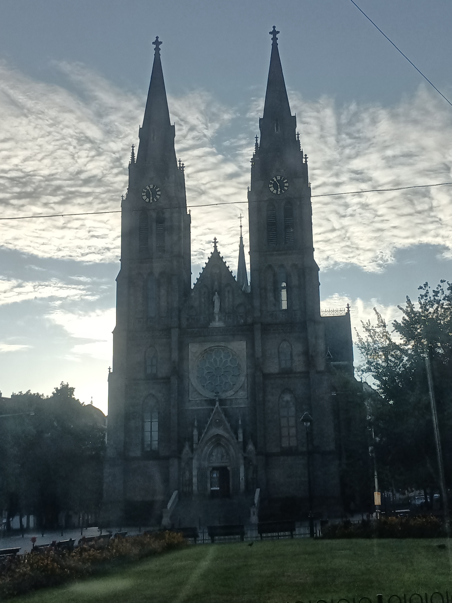 The image shows the Basilica of St. Peter and St. Paul in Vyšehrad, Prague, Czechia. The gothic basilica features two prominent spires, each topped with a cross. A large clock face is visible on each spire. In the center of the facade, a rose window is situated above the main entrance. The building is constructed from dark stone. The sky is partly cloudy with a soft, diffused light. In the foreground, a green lawn is visible, with a flowerbed and benches in the middle distance. Trees line the sides of the basilica and extend into the background. A power line runs horizontally across the upper portion of the image.