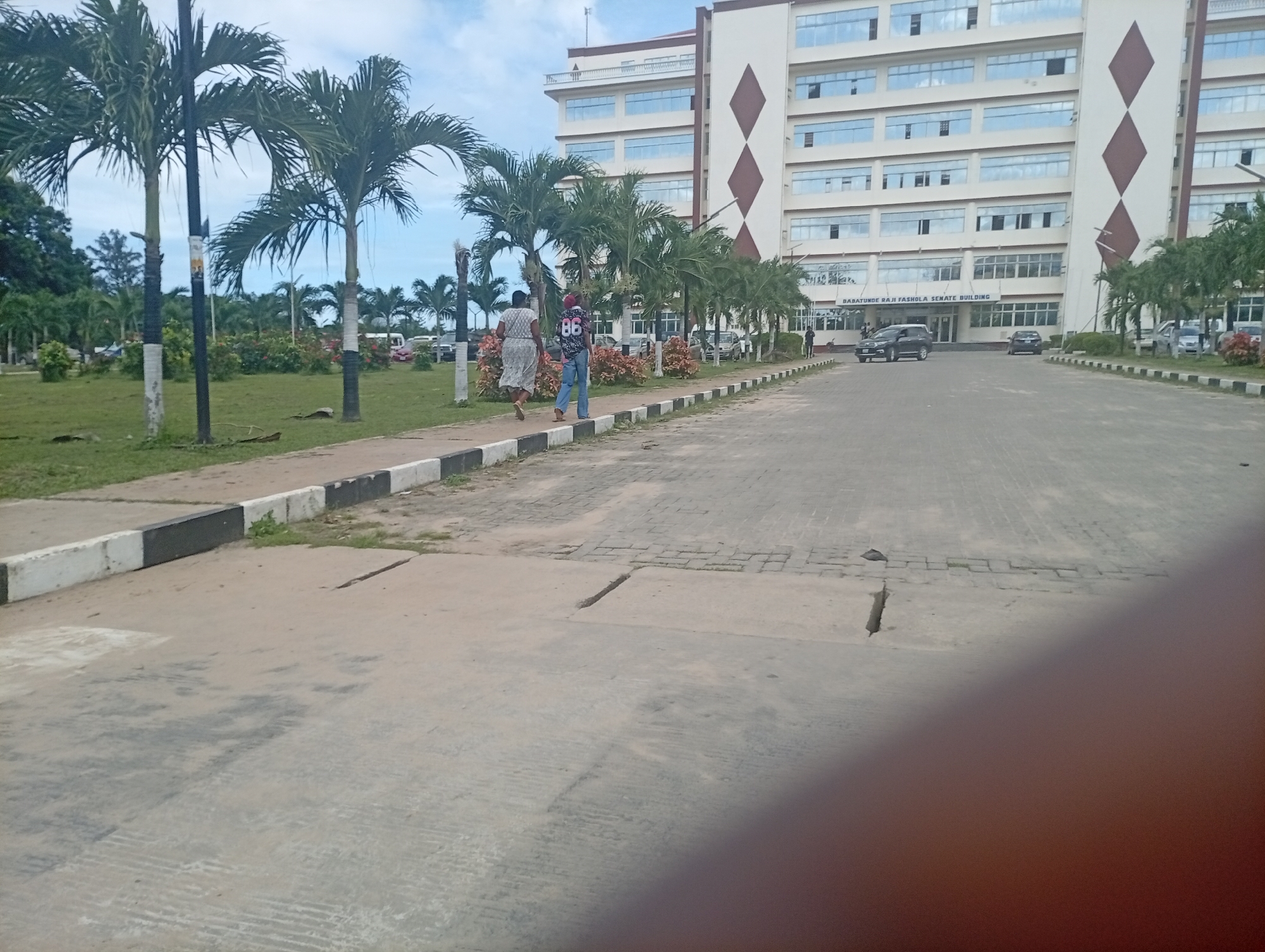 The image shows the Babatunde Raji Fashola Senate Building at Lagos State University (LASU) in Ijanikin, Nigeria. Two people are walking along a sidewalk towards the building. The man is wearing a shirt with the number 86 on it, and jeans. The woman is wearing a dress. There are palm trees and a lawn to the left of the sidewalk. Cars are parked near the building. The sky is cloudy.