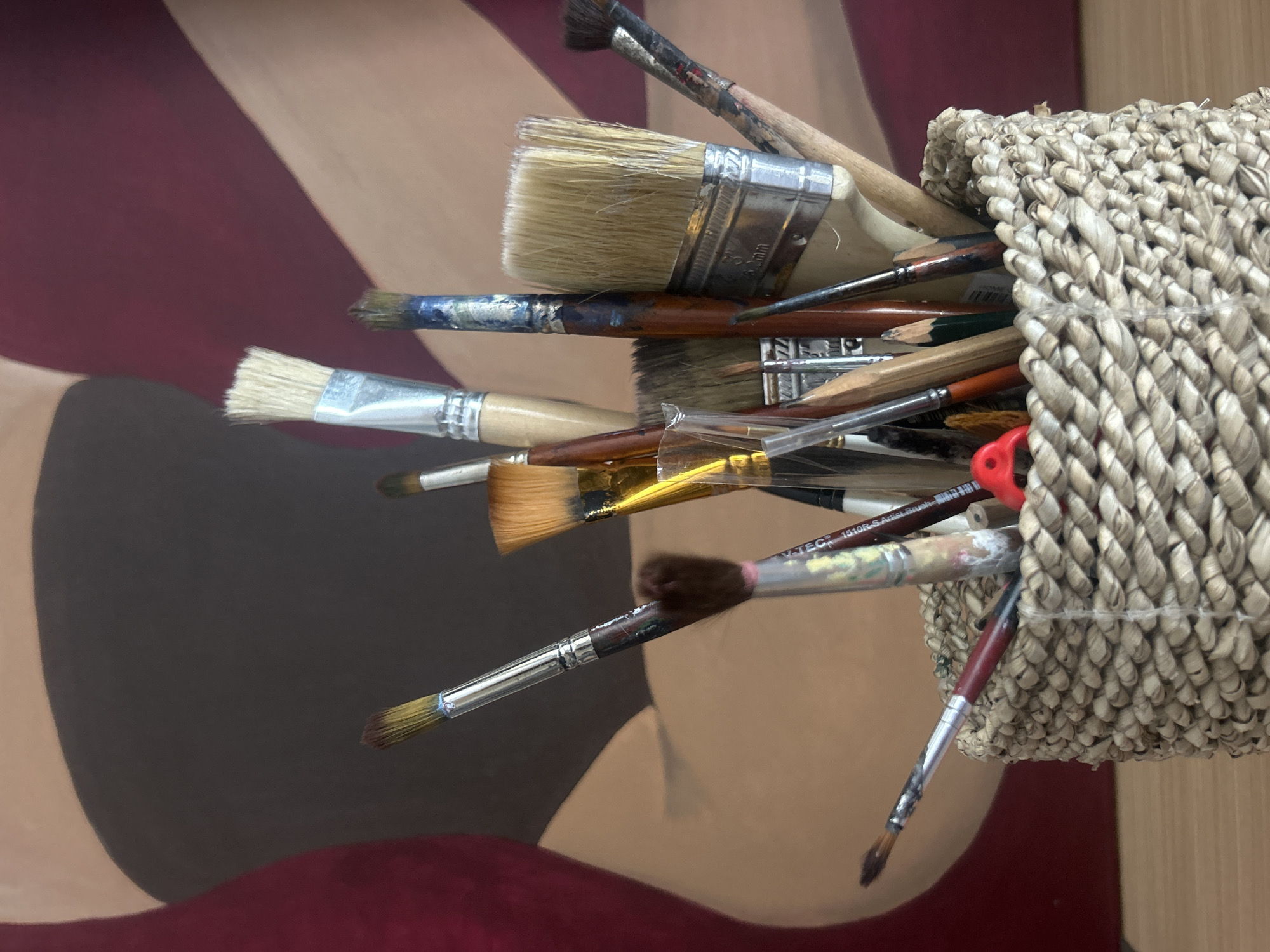 A close-up shot features a collection of paintbrushes and some pencils clustered within a woven basket. The basket appears to be constructed from a light-colored, fibrous material. The paintbrushes are of various sizes and styles, showing signs of use with dried paint on their bristles and handles. The basket is positioned against a backdrop of an artwork with broad strokes of maroon, brown, and tan, suggesting a contemporary or abstract style. The setting is probably located in a painter's studio. The precise city location in North Kuta, Indonesia, cannot be determined from this image.