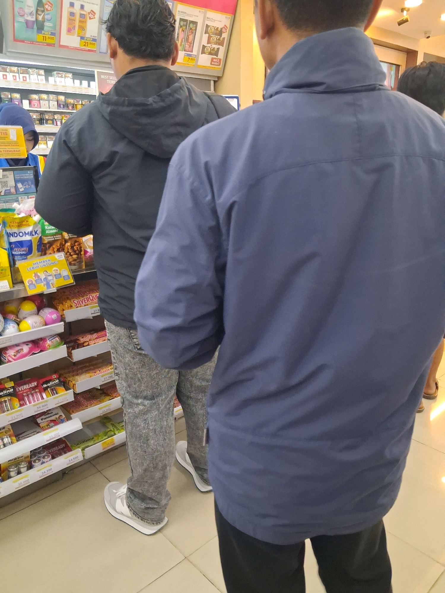 A low-angle, medium shot captures two individuals from behind in a brightly lit convenience store aisle in Surakarta, Indonesia. The person on the left wears a black hooded jacket and patterned gray pants, their back to the camera as they appear to be looking at products on a shelf. Their feet are clad in white sneakers with gray accents. To the right, a second individual is seen from the back, wearing a dark blue jacket and black pants. This person is positioned slightly closer to the camera, their shoulder obscuring some of the first person's torso.
Shelves laden with various products, including packaged snacks, drinks, and personal care items, are visible in the background. Notably, a yellow promotional sign with Indonesian text is attached to one of the shelves. The flooring appears to be a light-colored tile. The overall impression is of a mundane shopping scene within a typical Indonesian convenience store.