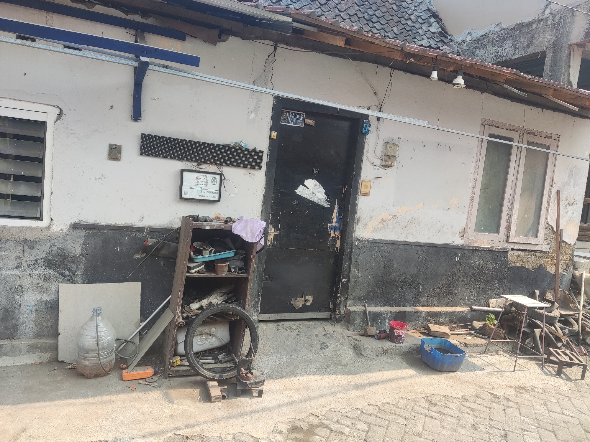 The image depicts a weathered building facade in Trihanggo, Indonesia. The building has a light-colored, cracked plaster exterior with a dark, distressed band running along its base. A black, weathered door is centrally located, marked with graffiti. To the left of the door, a window with horizontal slats is visible. Above the door, a sign with the numbers "113" is mounted. A white, metal awning extends horizontally across the facade.
In front of the building, a rough concrete area is scattered with various objects. To the left, a large, clear plastic water jug sits on the ground next to a pale, square panel. A wooden shelf unit is placed near the door, filled with miscellaneous items including tools, a purple cloth, and a bicycle wheel. Further to the right, several discarded tires are piled near a metal table with a worn top. A blue plastic tub filled with water is also present on the ground. The environment suggests an outdoor, utilitarian space, possibly a workshop or storage area, with a tiled pavement in the foreground. The overall impression is one of disrepair and functional clutter.