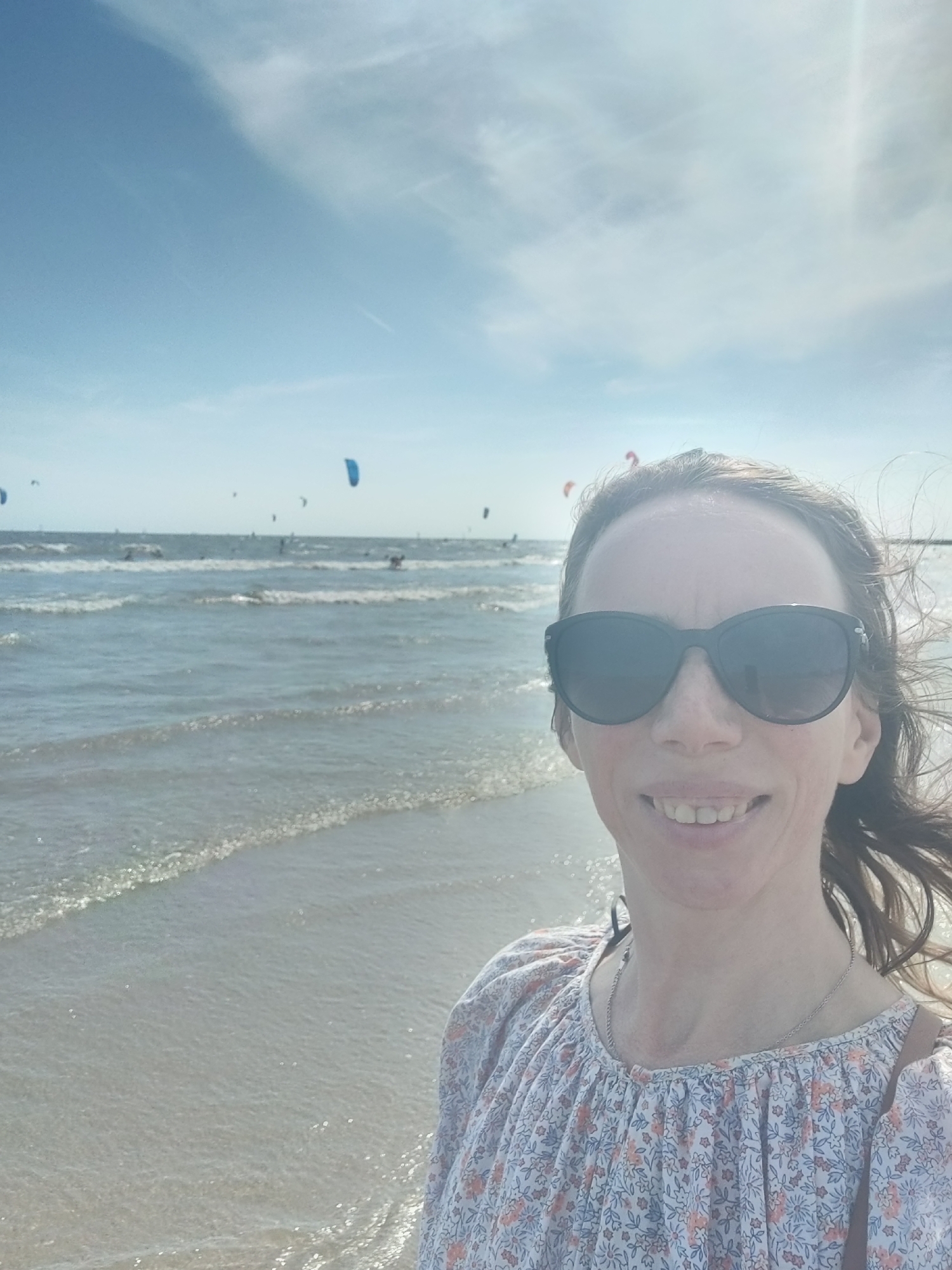 A woman wearing sunglasses and a floral-patterned shirt stands on a beach, taking a selfie. In the background, the sea is visible with waves and several kitesurfers in the distance. The sky is blue with scattered clouds, and the sun is shining brightly, creating glare on the water. The scene appears to be at Scheveningen beach in The Hague, Netherlands, given the presence of kitesurfers which is a popular activity at this location.