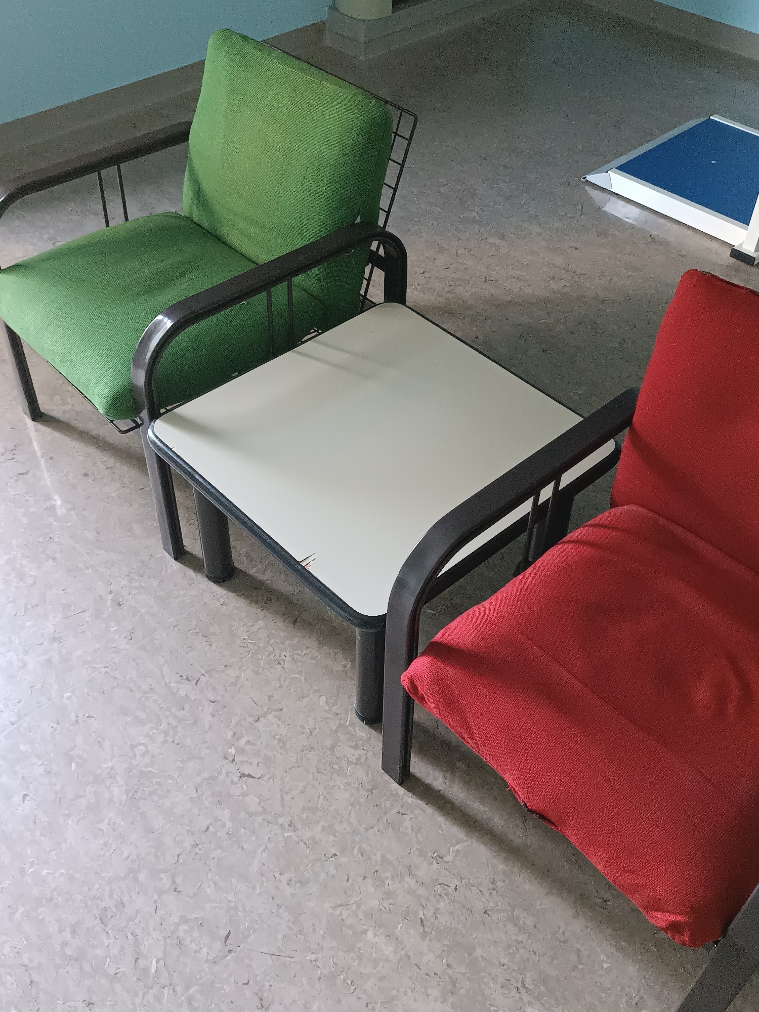 The image shows an indoor scene featuring two chairs and a small square table. One chair is upholstered in green fabric and has a dark metal frame. The other chair, partially visible on the right side of the image, is upholstered in red fabric and also has a dark metal frame. Between the chairs is a square table with a light-colored top and dark metal legs. The flooring appears to be a light-colored tile with a marbled pattern. In the background, a light blue wall is visible, and a rectangular object with a blue surface and white trim is placed on the floor. No people are present in the image.