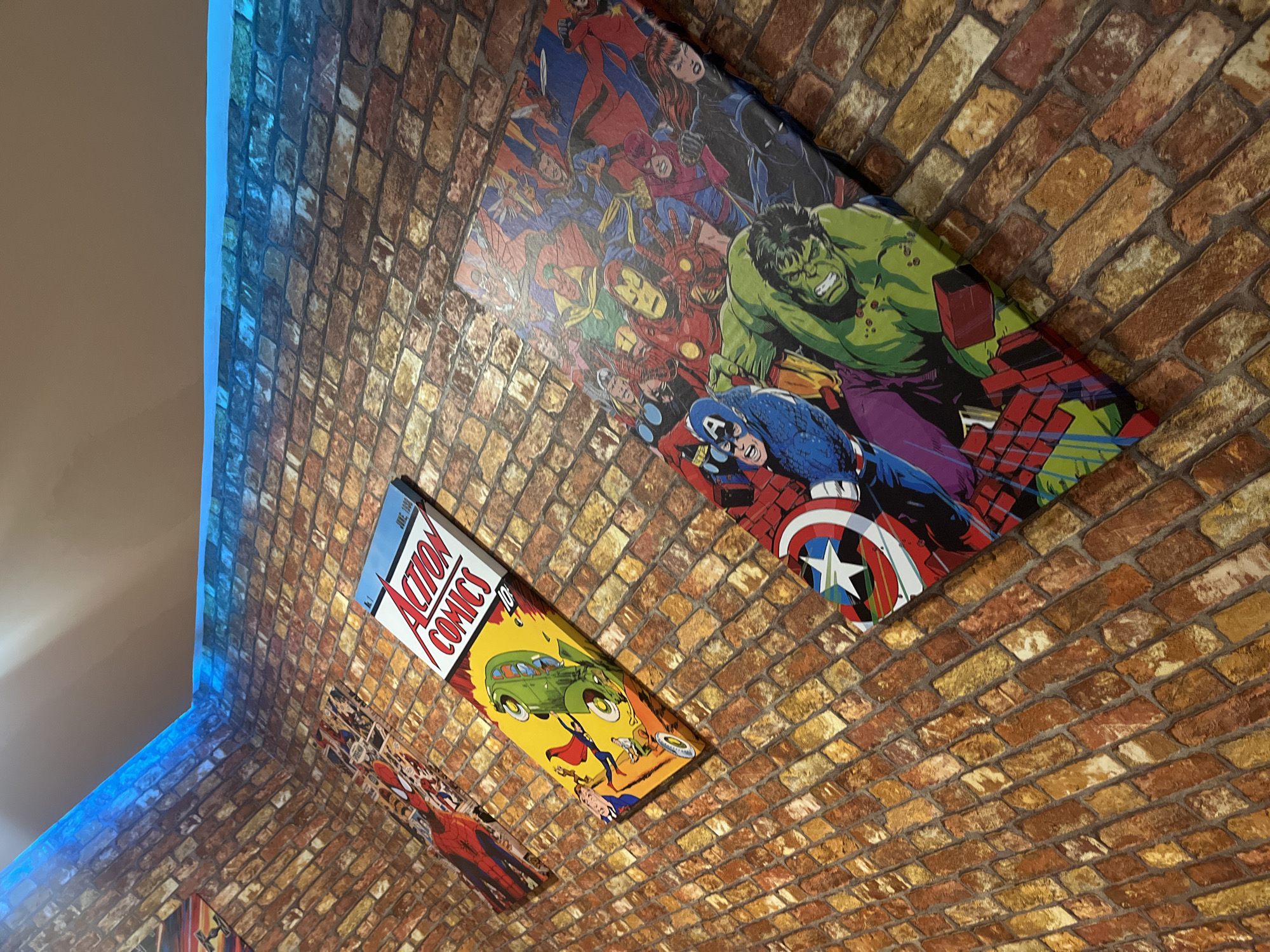 The image shows a portion of a brick-patterned wall, likely indoors, adorned with comic book-themed artwork. The artwork includes depictions of various superhero characters, such as Hulk, Captain America, and others, along with a large "Action Comics" cover. The photograph's angle suggests it was taken from below, looking upward and across the wall's surface. The scene includes a blue-lit edge, likely part of the ceiling or wall structure, which contrasts with the warm tones of the brick pattern and artwork. There are no people visible in the image. The location is not discernible from visual information, and the city of Porlamar, Venezuela, cannot be definitively determined.