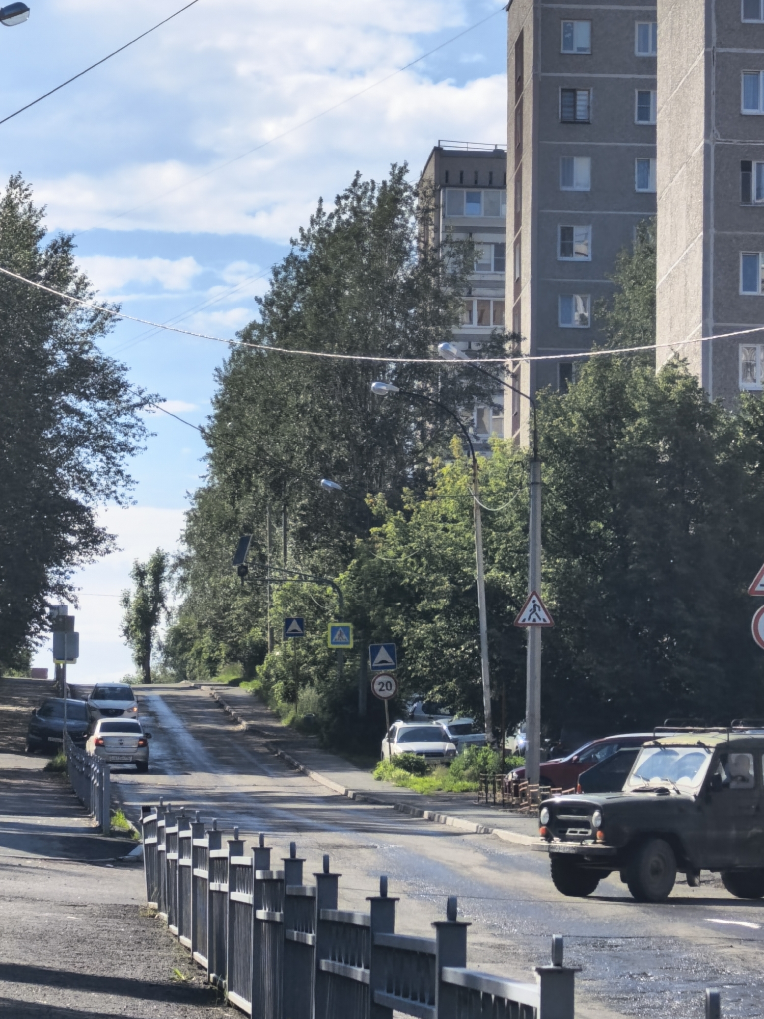 This media file shows a street scene on a sunny day. The scene is dominated by a road that slopes upwards and is lined with tall trees on either side. In the background, there are several multi-story apartment buildings, characteristic of urban or suburban environments. The sky is blue with some white clouds, suggesting fair weather.
Several cars are visible on the road. In the foreground, a dark green off-road vehicle is parked or moving slowly. Further up the road, a white car is driving uphill, followed by other vehicles. There are also traffic signs visible, including pedestrian crossing signs and a speed limit sign indicating "20". The road surface appears wet in places, possibly from recent rain or cleaning.
The overall impression is one of a typical day in a residential area with some traffic. The time of day appears to be daytime, given the bright sunlight casting shadows. There are no people prominently featured in the image, with the focus being on the vehicles, buildings, and the street environment. The scene conveys a sense of everyday life in a built-up area.