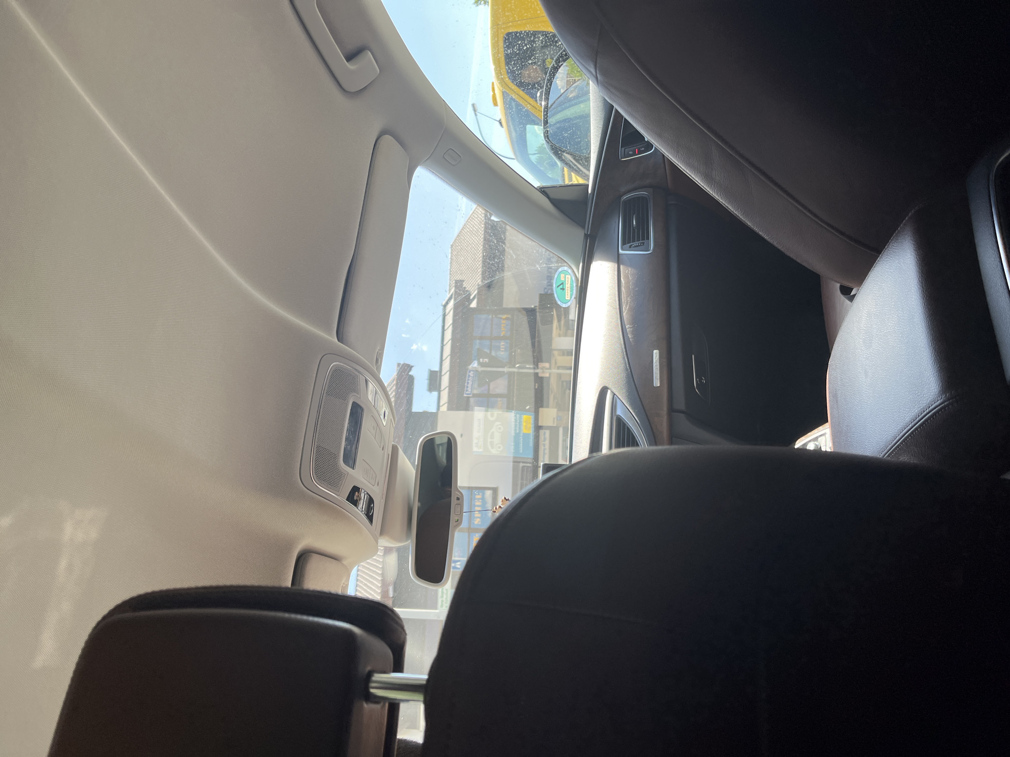 The image provides an interior view from the back seat of a car, looking forward through the windshield. The scene is set in Garmisch-Partenkirchen, Germany, where the car is parked or stopped. The car interior features dark brown leather seats, a wood-grain dashboard, and a sunroof control panel. Outside the car, a yellow taxi cab is visible, along with buildings and trees. The lighting suggests it is daytime, with bright sunlight.