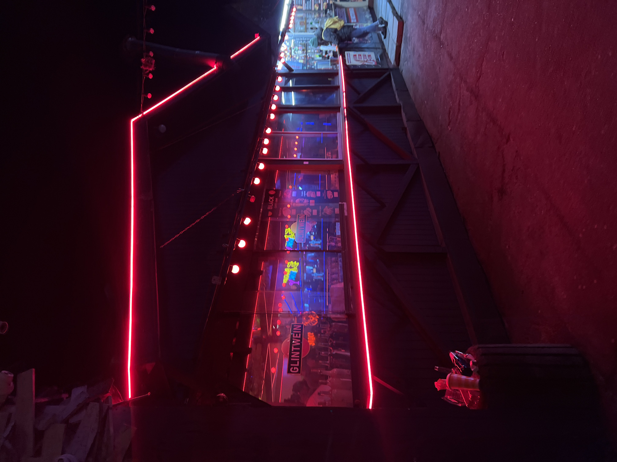 The image shows a street scene in Odesa, Ukraine. A building is illuminated with red neon lights, outlining a rectangular structure. A sign with the word "GLINTWEIN" is visible. The scene is dimly lit, and the red neon lights create a strong contrast against the dark background. On top, there is a person.