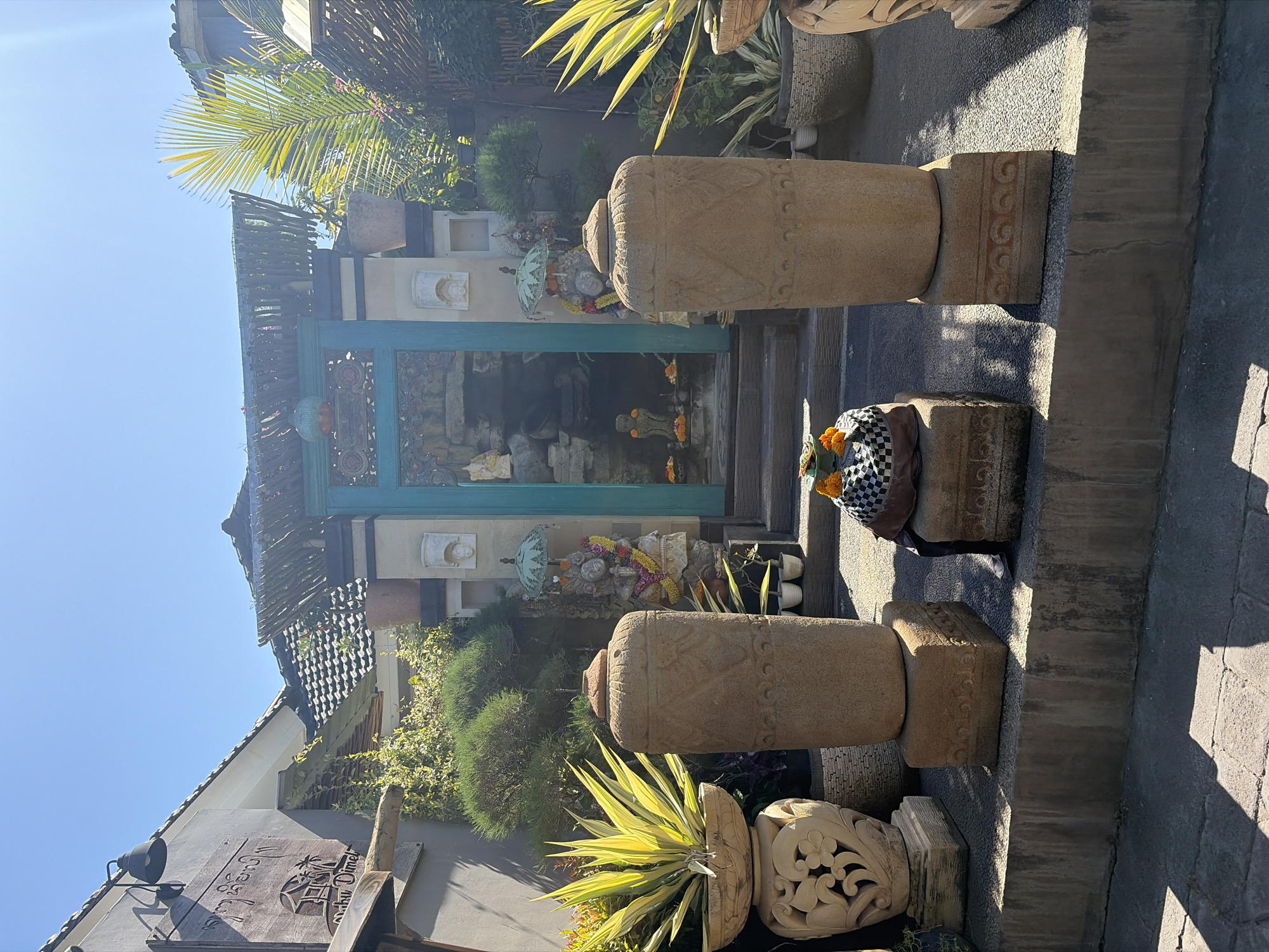 A frontal, eye-level, medium shot captures an outdoor scene featuring a building entrance and surrounding decorative elements, likely in Kuta Selatan, Indonesia. The entrance is framed by a teal-colored, ornately carved wooden doorway. Above the doorway, a rustic roof structure made of woven materials and bamboo is visible.
In front of the entrance, two large, cylindrical stone pillars with carved patterns stand on square bases. A smaller, square stone pedestal between them holds a checkered fabric bundle adorned with orange flowers. To the left of the entrance, a large, ornate stone planter contains a spiky, yellow-green plant. To the right, a similar stone planter is partially visible.
Behind the entrance, a rock garden with water features and small statues can be glimpsed through the doorway. Statues, possibly religious figures adorned with floral garlands, are placed on either side of the entrance. Lush green foliage and trees surround the area. The ground is paved with dark gray stones, and shadows from the pillars and foliage are cast across it, indicating a bright, sunny day with the sun positioned high in the sky, as suggested by the clear blue sky. The architecture and ornamentation suggest a Balinese or Southeast Asian aesthetic.