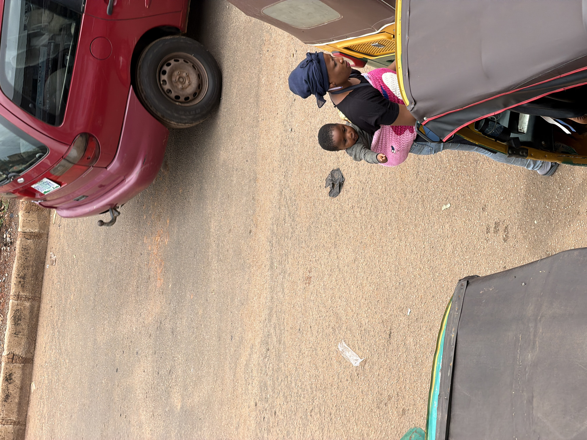 This image captures a street scene in Bukuru, Nigeria. In the foreground, a red car is parked on a paved road. Its rear wheel and part of its bumper are visible. To the right of the car, a yellow and gray motorized tricycle, commonly known as an "okada" or "keke napep," is also present.
Inside the tricycle, a woman wearing a dark headscarf and a black top is seated, holding a baby or young child. The child is wrapped in a pink and white knitted material, possibly a blanket or sling. The woman appears to be looking upwards or to the side, while the child is looking towards the viewer. The tricycle has a dark gray canopy.
On the road, next to the tricycle, a dark object, possibly a piece of clothing or a bag, lies on the ground. Further to the right, a small piece of clear plastic is also visible on the road surface. The overall scene suggests an everyday street environment in Nigeria, likely during daylight hours given the lighting conditions. No specific time of day or weather conditions are explicitly indicated, though the paved road and clear visibility suggest fair weather. There is no visible text in the image.
