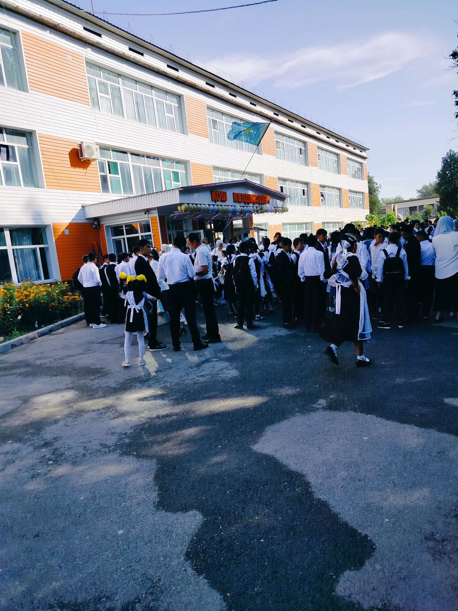 The image shows a group of students in uniform standing outside a multi-story building. The building has an orange and white facade with numerous windows. A flag, possibly the flag of Kazakhstan, is visible on the top left of the building. Several students are dressed in black and white uniforms, with some wearing bows and ribbons, suggesting a formal occasion or the beginning of the school year. There are also adults present, some in formal attire. The ground appears to be asphalt, with some reflections from what might be puddles or a wet surface. There are trees and other greenery visible in the background. The scene appears to be in an outdoor public space, likely a school campus. The text on the building archway, "КОЛ КЕНТАУ", indicates the location is Kolkentu, Kazakhstan.