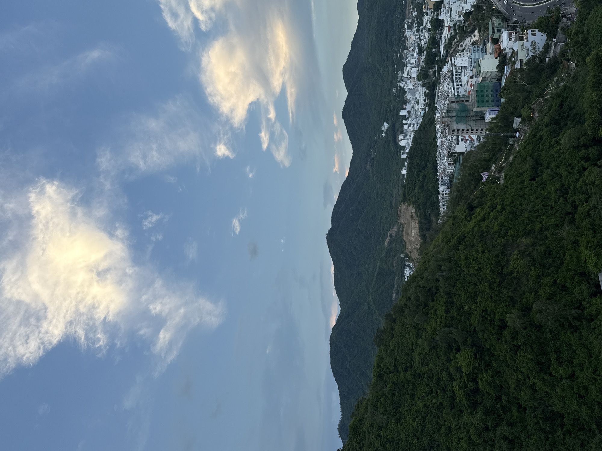 The image shows a panoramic view of a coastal city built on steep hillsides, likely in Nha Trang, Vietnam, given the location context. The scene is captured during what appears to be either sunrise or sunset, as indicated by the golden hues in the clouds and the soft lighting on the landscape. The sky is a blend of blue and soft grey, with scattered, fluffy clouds, some illuminated by the sun.
The foreground is dominated by a dense, dark green hillside covered in lush vegetation, suggesting a mountainous or hilly terrain. Descending from the crest of the hill is a sprawling urban area with numerous buildings, predominantly white and light-colored, clustered closely together. Some structures appear to be in various stages of construction, with exposed scaffolding visible on a few larger buildings. The buildings are built along the contours of the hills, creating a tiered effect.
In the distance, more mountains, also covered in vegetation, can be seen. A road with some vehicular traffic is visible winding through the city, indicating it's a populated and active area. The overall impression is one of a vibrant city nestled harmoniously within a dramatic natural landscape. There are no visible people or distinct objects that stand out, the focus being on the expansive scene. The time of day implies a potentially peaceful atmosphere, with the fading or rising sun casting a warm glow over the city and its surroundings. The weather appears clear with some cloud cover.