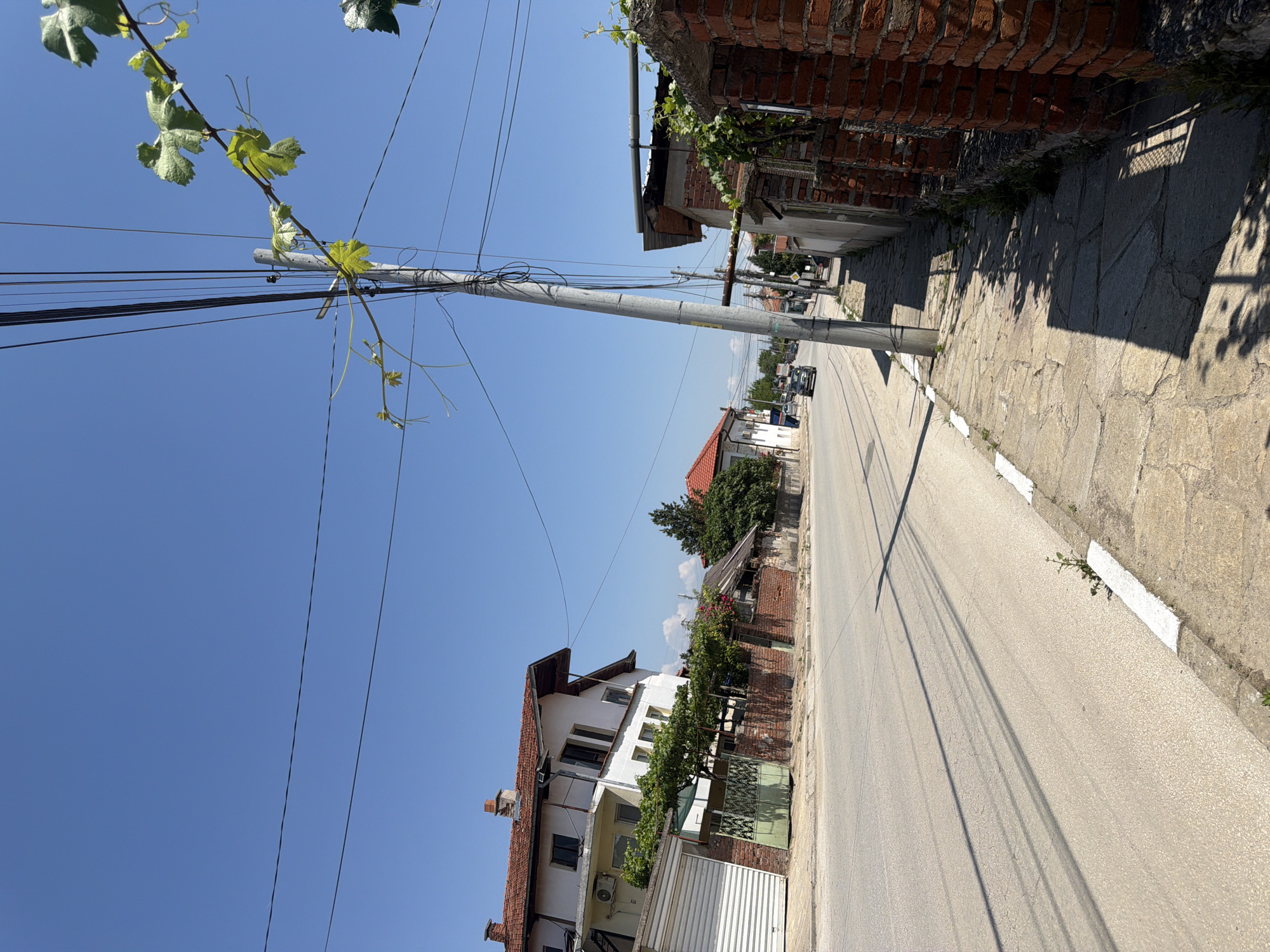 The image depicts a street scene in Zlatitrap, Bulgaria, on a clear, sunny day. The foreground is dominated by a paved sidewalk and an asphalt road. A tall, gray utility pole stands on the right side of the road, laden with numerous wires. To the right of the pole, a red brick wall runs along the sidewalk, partially overgrown with green vines. Across the street, on the left side, a multi-story white building with a red tiled roof is visible, featuring a white garage door in front of it. A green metal fence separates the building from the sidewalk. Further down the street, several other buildings with red tiled roofs and trees can be seen. A few cars are driving on the road in the distance. The sky is a vibrant, cloudless blue.