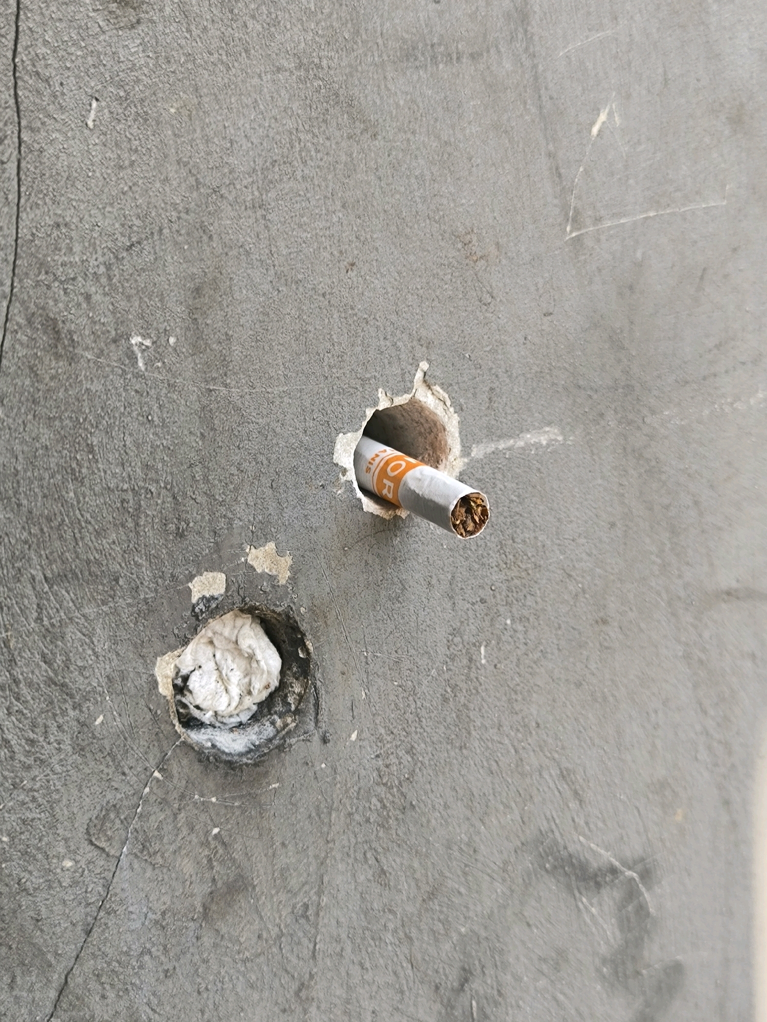 A close-up shot captures a gray, textured concrete surface. Two distinct circular holes are visible. In the upper right of the image, a cigarette is inserted into a hole, with its orange and white packaging partially visible, and the tobacco end exposed. The lower left hole is filled with a wad of white material, possibly paper or cotton, secured with some dark adhesive. The surface of the concrete is scratched and worn, suggesting an outdoor urban environment.