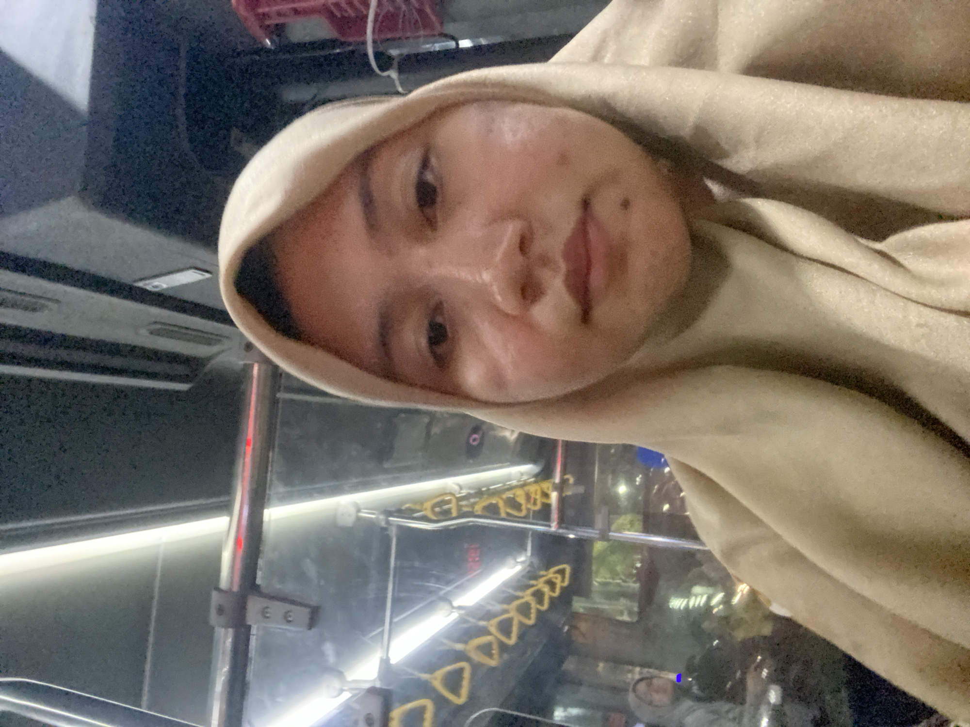 A close-up, selfie-style shot shows a young woman wearing a light brown hijab. She is positioned in the foreground, with her face and upper body filling most of the frame. Her expression is neutral, and her gaze is directed towards the camera. The background appears to be the interior of a public transportation vehicle, likely a bus, given the presence of yellow handholds hanging from overhead rails and the general aesthetic of the interior. The lighting suggests it is either evening or nighttime, with artificial lights from within the vehicle and possibly outside illuminating the scene. A blurred reflection or image of another person wearing a grey hooded jacket can be seen in the background, suggesting other passengers are present. The location is identified as Banjarmasin, Indonesia.