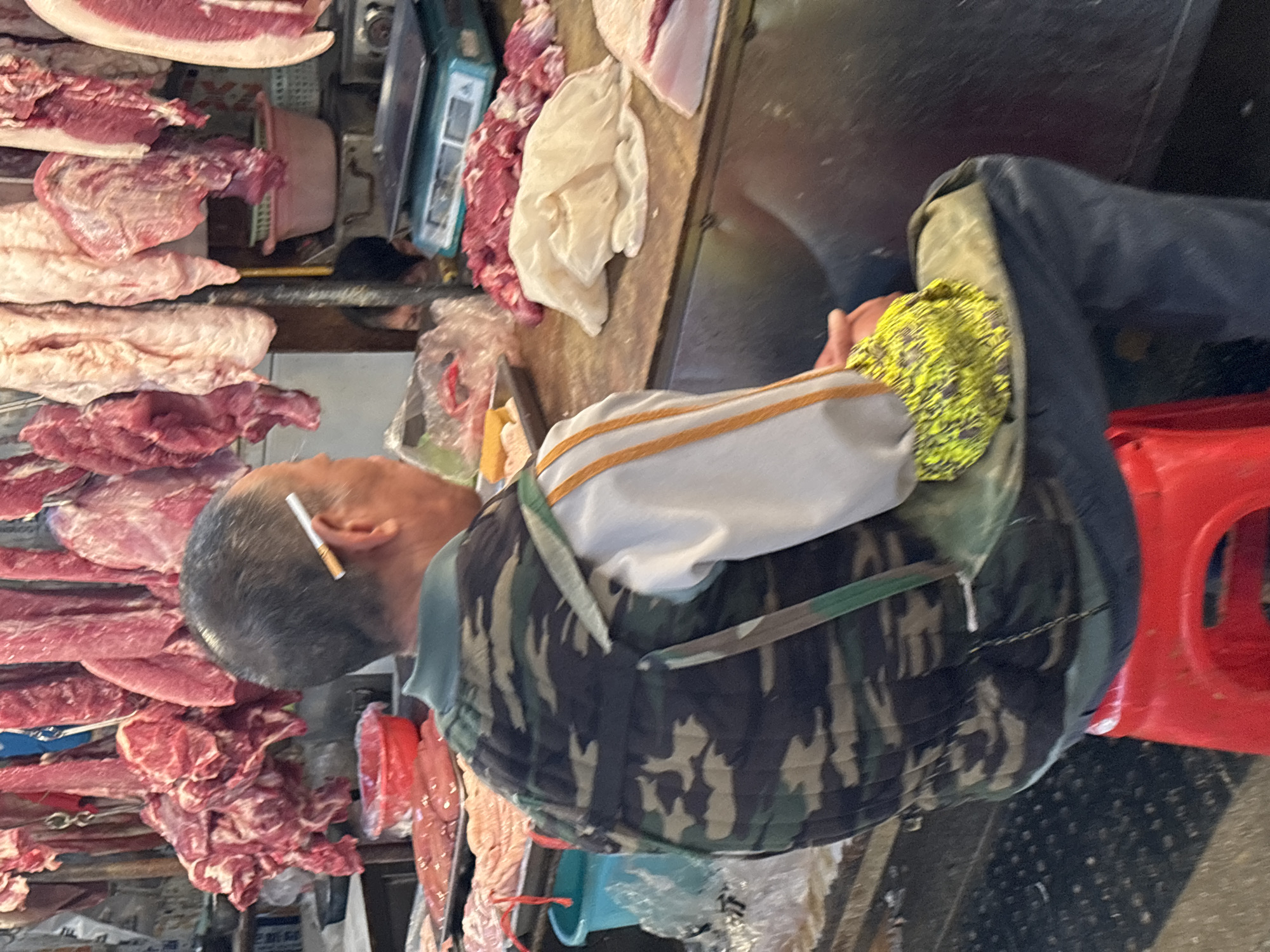 An adult male with a cigarette behind his ear sits near raw meat on display, likely in a market. The location is believed to be in Jiangjin, China, based on image context. The man is wearing a camouflage jacket. There is a red plastic stool to the side. The environment seems to be a commercial space.
