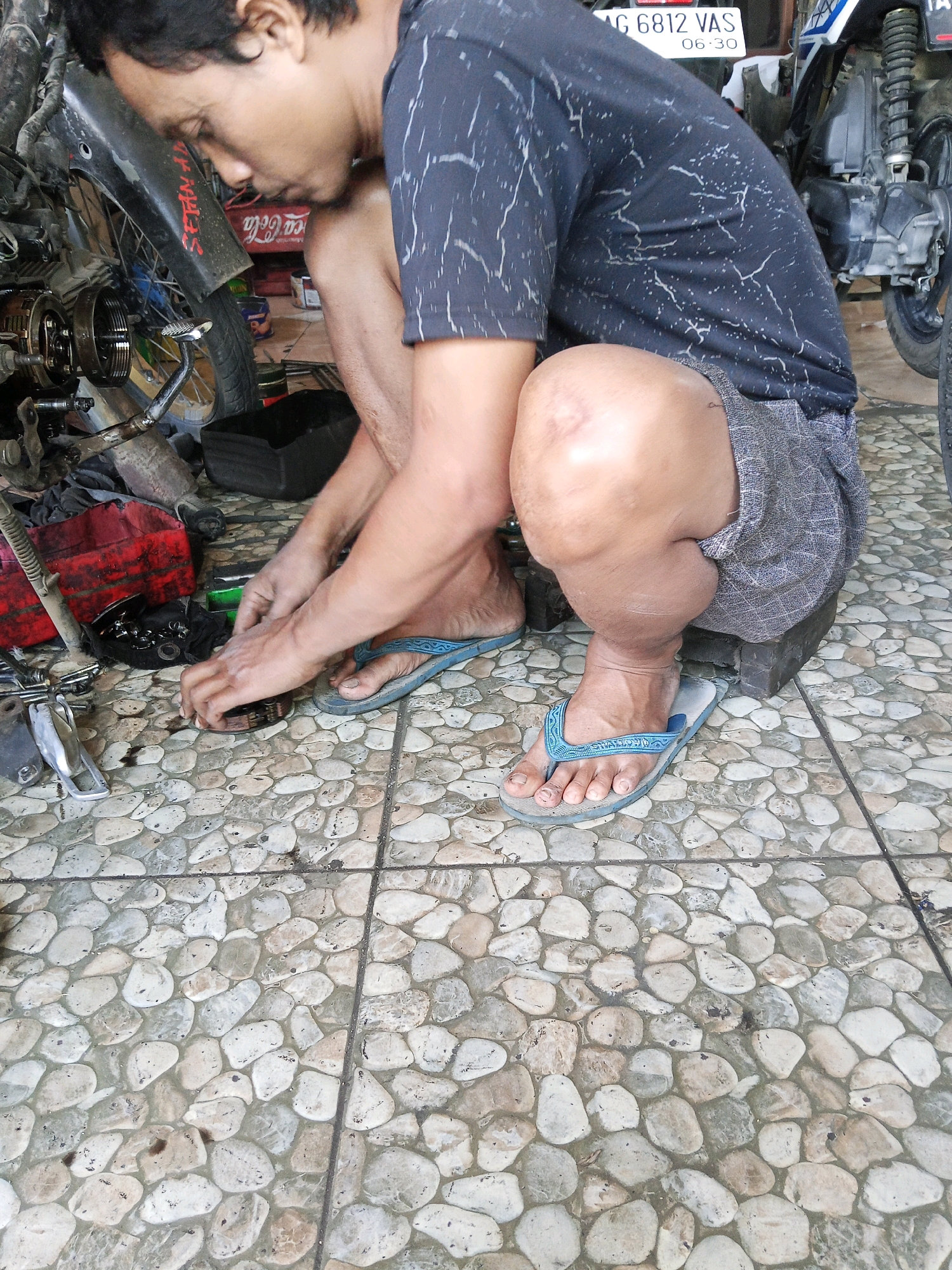 A man is kneeling on a stone-tiled floor, working on a motorcycle in an outdoor workshop in Wates, Indonesia. He is wearing a dark t-shirt with a white speckled pattern and patterned shorts. His right hand is visible, manipulating a small, circular metal object. His left hand is also engaged in the task. He is wearing blue flip-flops. To his left, a motorcycle engine component and a red plastic container are visible. Behind him, a motorcycle with a white license plate displaying "AG 6812 VAS 06.30" is present. The environment appears to be a shaded area, possibly an open-air repair shop or a residential courtyard.