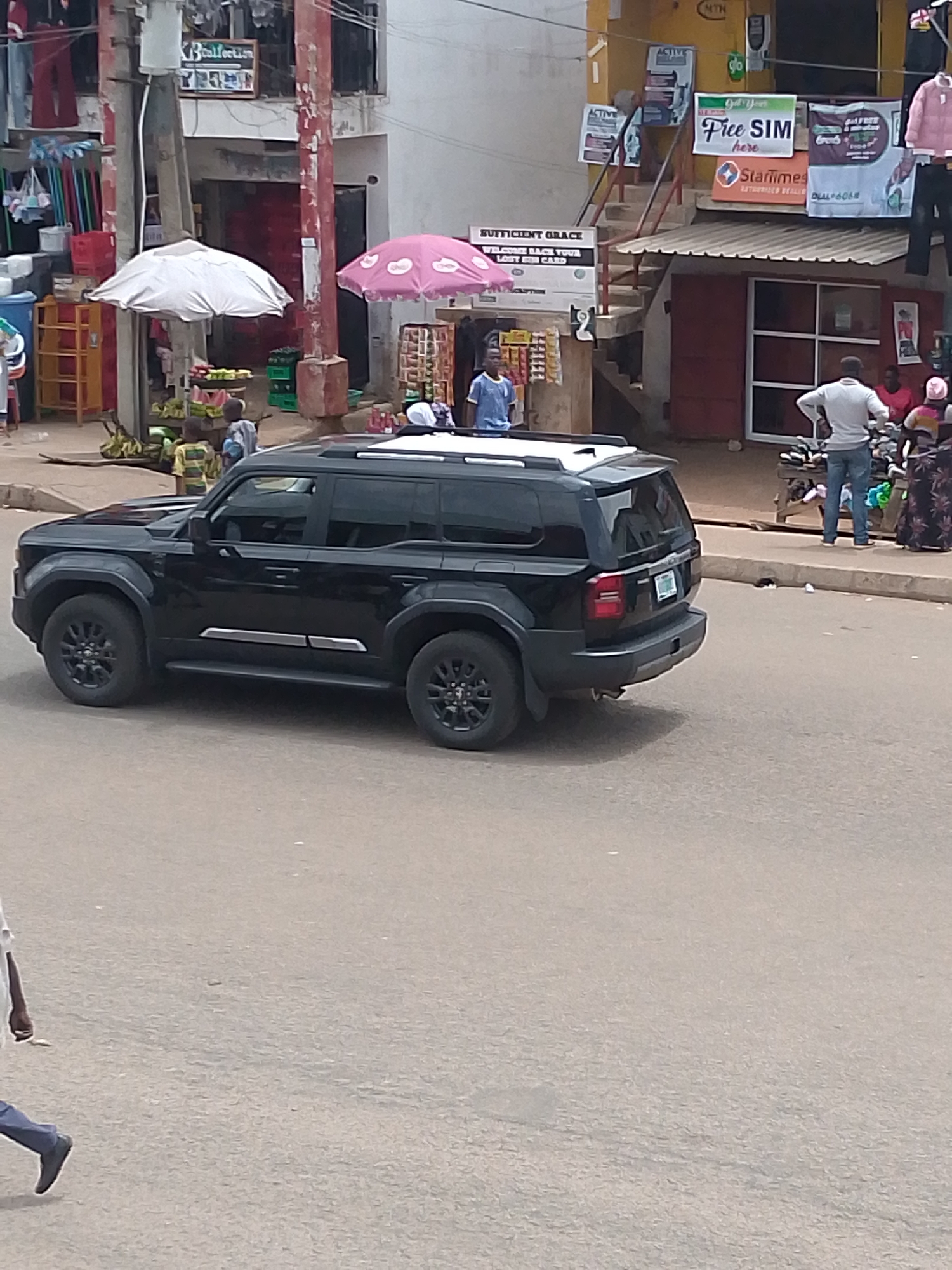 The image depicts a black SUV parked on a street in Bukuru, Nigeria. In the background, people are walking and standing near a building with various signs and advertisements, including one for "Free SIM." There are also umbrellas shading vendors selling goods, including bananas and other items, on the street.