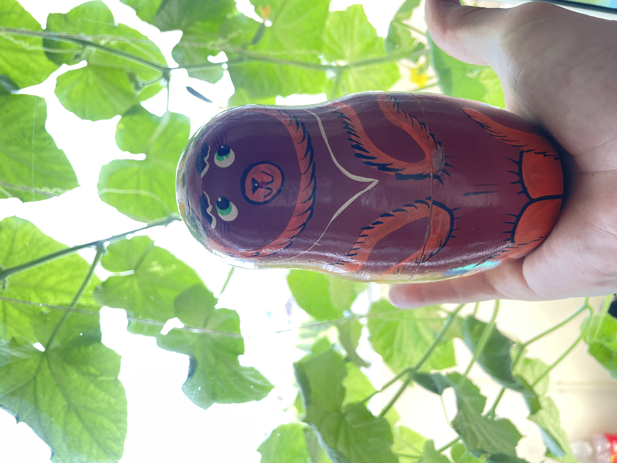A hand holds a wooden matryoshka doll in front of a window with green foliage. The doll is painted dark red with orange and black accents, depicting a bear's face with green eyes and black eyebrows on its upper half. Below the face, there are painted details resembling a chest and paws. The background consists of blurred green leaves and stems, illuminated by bright natural light filtering through the window. The overall scene appears to be indoors, near a window, with the focus on the doll and the hand holding it. The location of Laishevo, Russia is not discernible from the image.