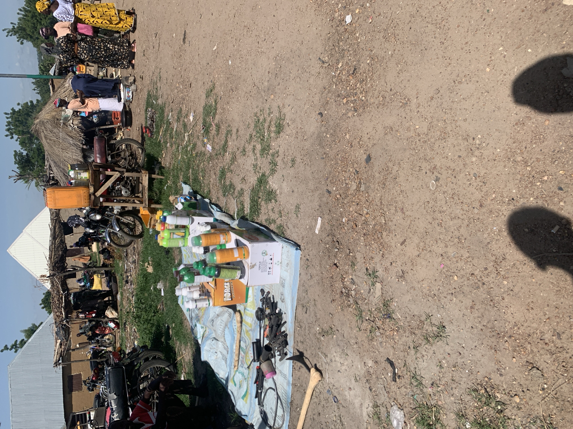 This image captures a busy outdoor market scene in Nigeria, likely during daylight hours under a clear sky. The foreground features a stall laid out on a patterned blue and white cloth, displaying an assortment of items. These include numerous bottles of various sizes and colors, many appearing to be cleaning products or beverages, alongside some cardboard boxes. Among the bottles, a box with the label "HORIZON OIL" is clearly visible, along with another box labeled "STYLE". Scattered on the cloth are also various tools, including what looks like a sickle and what appears to be a rifle or its components, suggesting a diverse range of goods for sale.
In the background, a collection of motorcycles is parked, indicating transportation is a significant aspect of the location. Behind the motorcycles, a makeshift structure with a thatched roof is visible, suggesting a semi-permanent market setup. Several people are present, some standing and interacting near the stall and motorcycles, while others are seen in the background, dressed in traditional Nigerian attire. The overall atmosphere appears lively, with the hustle and bustle typical of a local marketplace. The ground is a mixture of dirt and gravel, with sparse patches of grass, consistent with an outdoor setting in a rural or semi-urban area of Nigeria. The presence of motorcycles and the style of clothing suggest a contemporary setting.