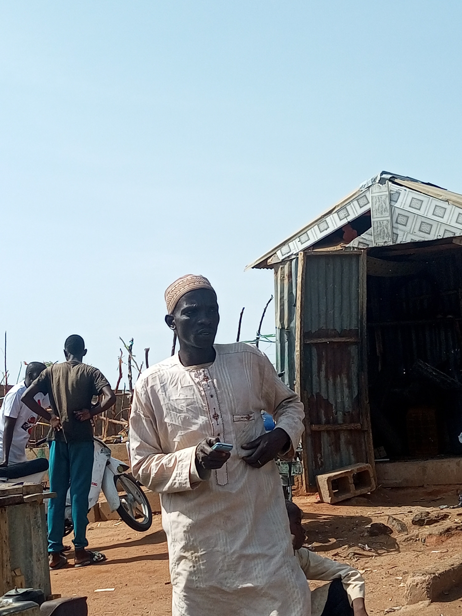 The image captures an outdoor scene, likely in Gombe, Nigeria, featuring several individuals and simple structures under a clear, bright sky.
In the foreground, a dark-skinned man stands, facing slightly right and looking off into the distance. He wears a light-colored traditional long garment (possibly a djellaba or similar tunic) with embroidered details on the chest and cuffs, and a patterned kufi cap. He holds a light blue mobile phone in his left hand.
Behind him, to the left, are two other men whose backs are largely turned to the camera. One wears a dark t-shirt and teal or blue pants, standing with his hands on his hips. The other, partially obscured, wears a white t-shirt. Behind these men, a white motorbike or scooter is parked on the dusty ground. Further back, sparse wooden sticks are visible, suggesting fencing or dry vegetation.
On the right side of the frame, a small, roughly constructed shack or shed is prominent. Its walls are made of corrugated metal, showing signs of rust and wear. The roof appears to be pieced together from various materials, including what looks like repurposed ceiling tiles with square patterns. The door of the shack is open, revealing a dark interior. Concrete blocks are placed on the ground near the shack. In the lower right corner, a child is partially visible, sitting on the ground.
The time of day appears to be midday or early afternoon, indicated by the strong sunlight and relatively short shadows cast on the dusty, reddish-brown ground. The sky is a uniform, pale blue, suggesting clear and sunny weather. No discernible text is visible in the image. The overall setting depicts a casual gathering in what appears to be a local or semi-rural environment.