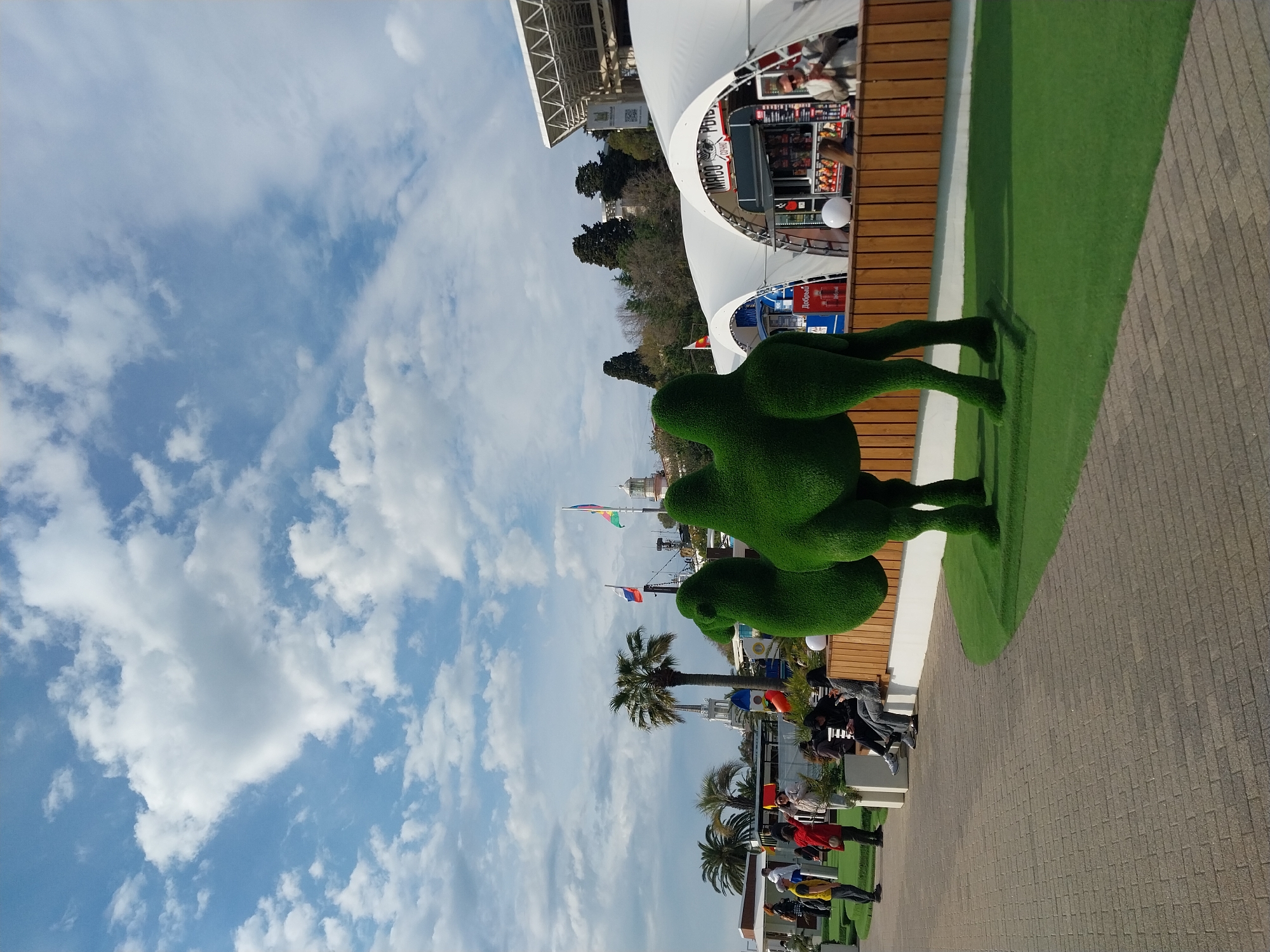 The image displays an outdoor leisure area, likely a promenade or park, under a bright, partially cloudy sky. In the foreground, two large, abstract green topiary figures stand on a strip of artificial green turf. This turf is bordered by a white strip, which separates it from a grey paved walkway constructed of interlocking bricks.
In the mid-ground, several people are present. Some are sitting on benches or chairs near kiosks, while others are standing. The kiosks feature white tent-like awnings and wooden facades. Visible text on one kiosk reads "Мясо Рыба" (Meat Fish), indicating food sales. Another red dispenser or cooler shows the brand name "Добрый" (Dobry) in Cyrillic. White spherical lights are mounted on posts or kiosk structures.
Further in the background, tall palm trees and other trees are visible, suggesting a warm climate. Flags can be seen flying, including a Russian tricolor flag and a rainbow-colored flag. An overhead white metal framework, possibly part of a larger structure or canopy, is also present. The scene is illuminated by clear daylight, casting soft shadows, indicating it is daytime. The overall impression is that of a relaxed, pleasant public space, consistent with a resort location like Sochi, Russia.