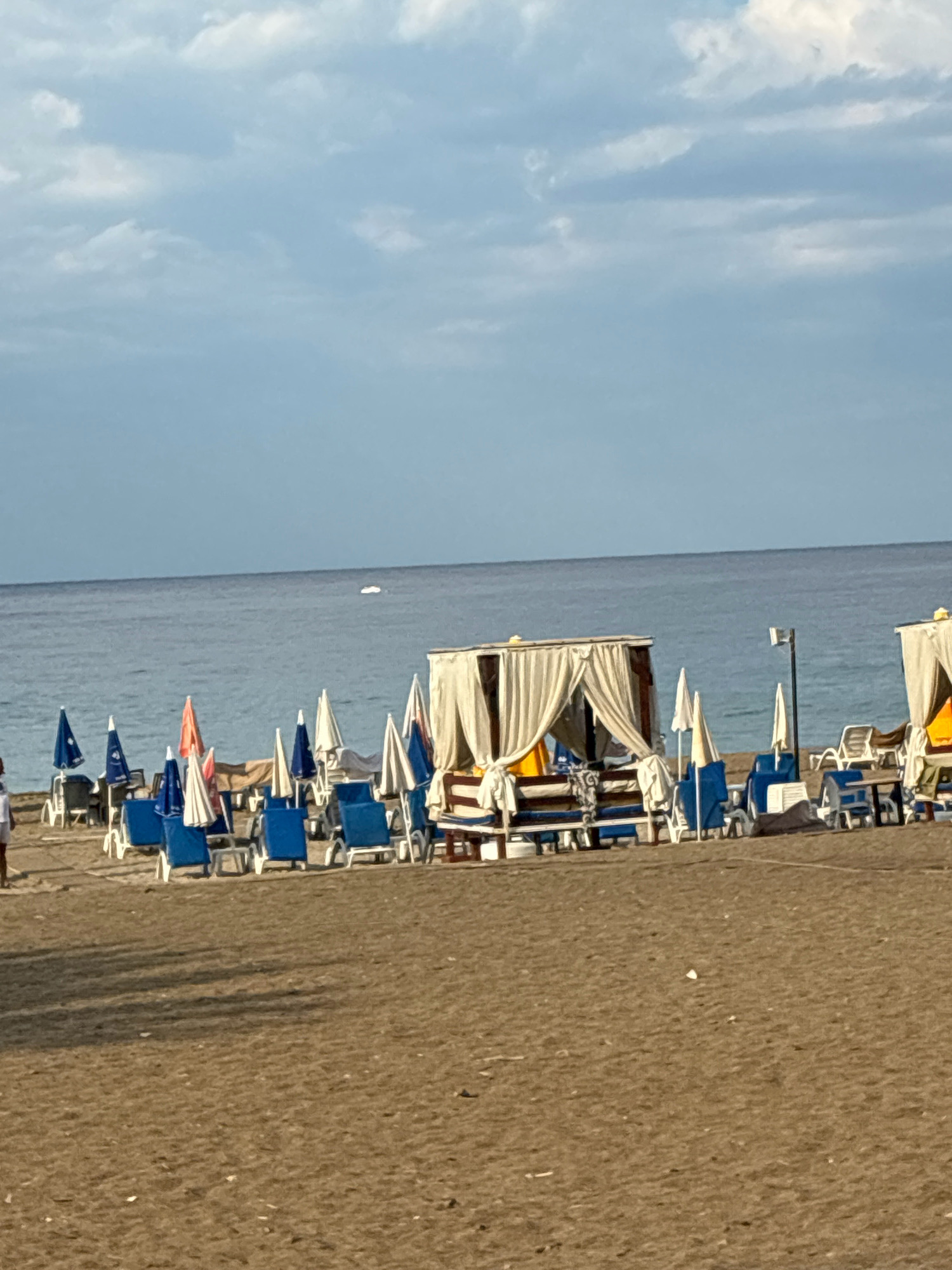 The image depicts a beach scene in Alanya, Turkey. In the foreground, there is a sandy beach with numerous lounge chairs and umbrellas arranged in rows. Several of the lounge chairs are blue and white, and some are accompanied by white umbrellas. In the middle ground, there are two cabanas with white curtains, offering shaded seating areas. One cabana is larger and more prominent, with a wooden structure and seating benches. The other cabana is smaller and appears to be part of a resort or service area. The sea is visible in the background, with a calm surface and a pale blue hue. The sky is partly cloudy, with patches of white and gray clouds against a light blue backdrop. A faint white object, possibly a buoy or a distant boat, is visible on the water. On the left side of the image, a person is walking away from the camera, dressed in casual clothing. The overall impression is a typical beach resort setting, ready for visitors to relax and enjoy the seaside.