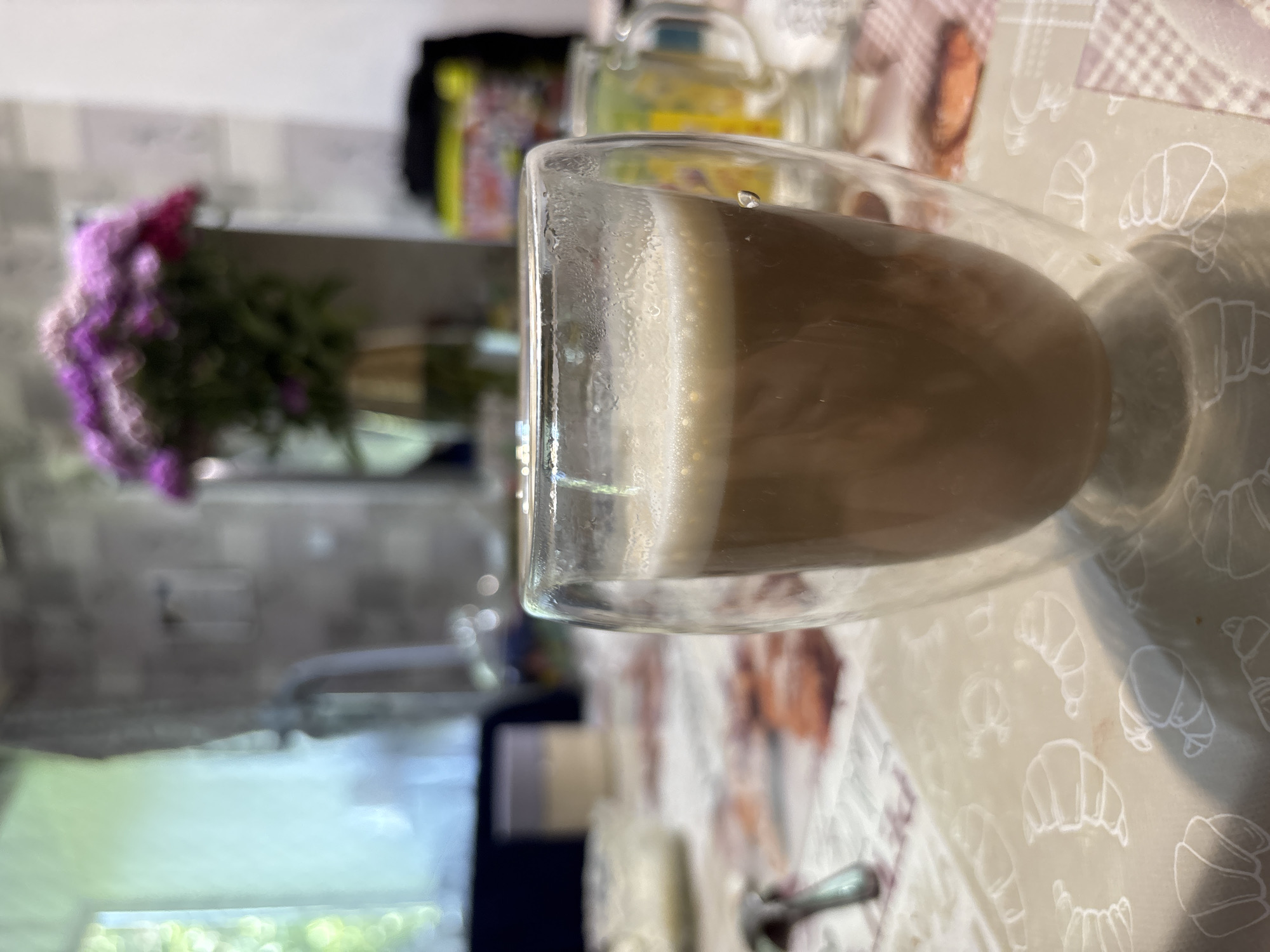 A clear glass mug with double walls, filled with a light brown beverage topped with a foamy head, is positioned on a table covered with a patterned tablecloth featuring illustrations of croissants. In the blurred background, a vase of purple flowers sits on a counter, and other objects such as jars and possibly food items are visible. The scene appears to be indoors, likely a kitchen or dining area. The specific location of Zubov, Russia, is not visually evident from the image itself, nor are there any people or actions depicted.