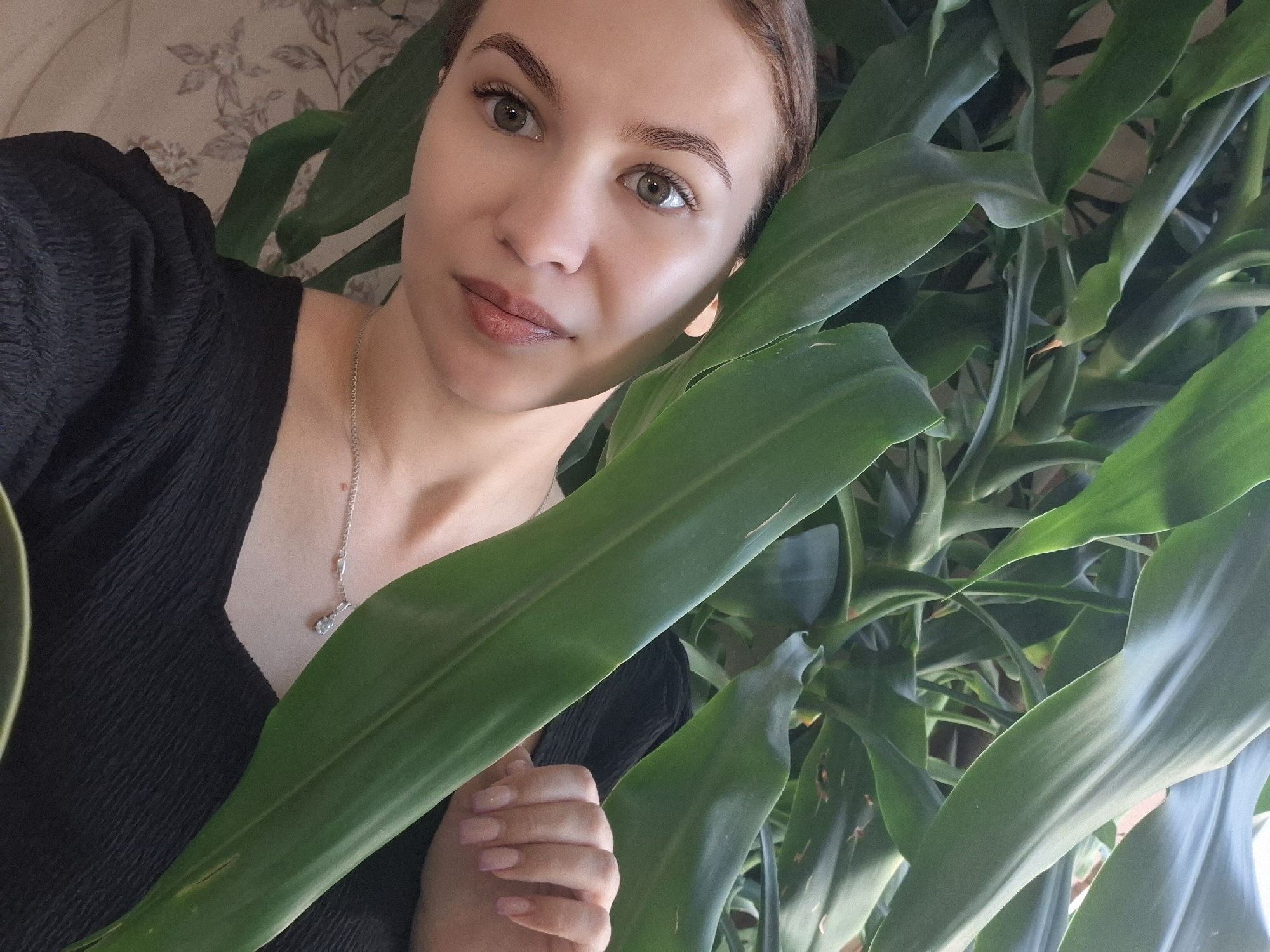 A young woman with fair skin and brown hair pulled back into a bun poses for a selfie. She wears a black top with short, puffed sleeves and a silver necklace with a pendant. The background is filled with the lush green foliage of a houseplant, obscuring a wallpapered wall in the upper left corner. Her hand is visible holding a leaf. The location is Nizhnyaya Gar', Russia, as inferred from the original query.