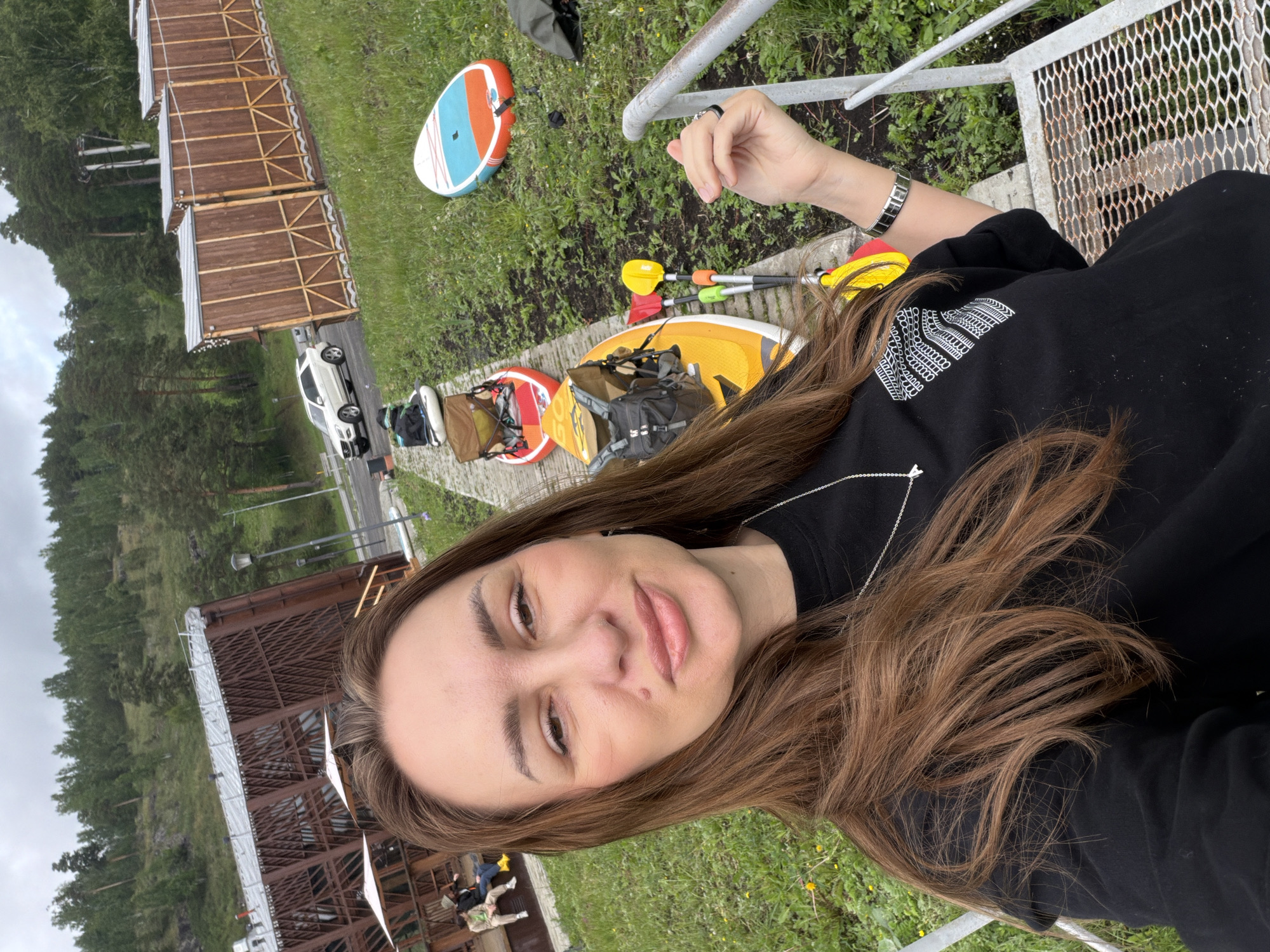 A woman takes a selfie outdoors in Mezhevoy, Russia, with several paddleboards lined up behind her. She is wearing a black sweatshirt and a silver necklace. The paddleboards are on a paved path next to a grassy area. In the background, there are wooden structures, trees, and a white car. The weather appears to be overcast.