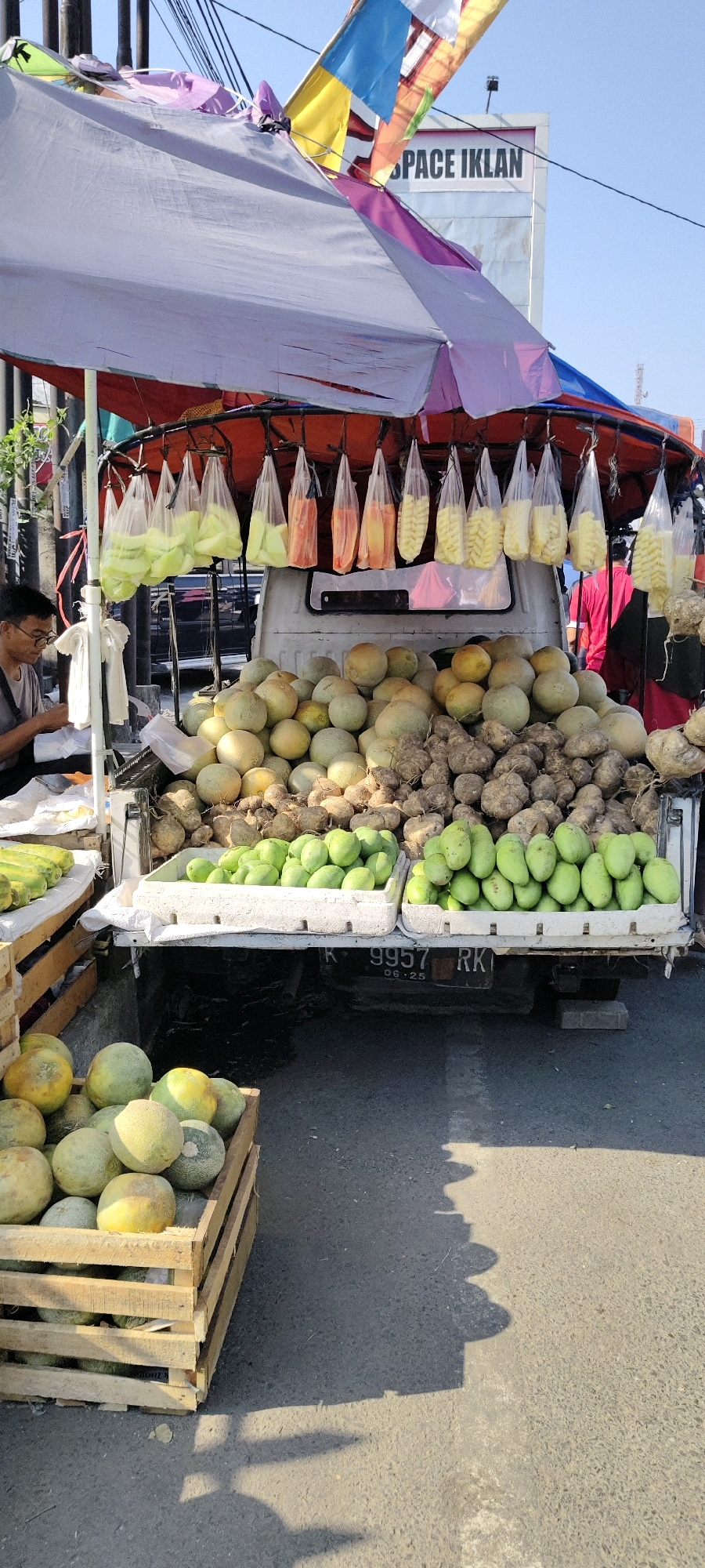 This image depicts an outdoor fruit stand in Kemadu, Indonesia, possibly along a roadside, bathed in bright sunlight. A white truck serves as the base for the stall, which is covered by a large, multi-colored umbrella. Suspended from the stall are plastic bags filled with sliced fruits, including what appear to be cantaloupe, guava, and pineapple. Piles of melons and root vegetables, possibly yams or jicama, are displayed in crates and directly on the truck bed. In the foreground, a wooden crate overflows with melons. A young man wearing glasses sits at the stall, appearing to be bagging produce. The background shows a clear blue sky, overhead wires, and a billboard with the text "SPACE IKLAN." The ground is paved, with shadows cast by the stall and its merchandise.