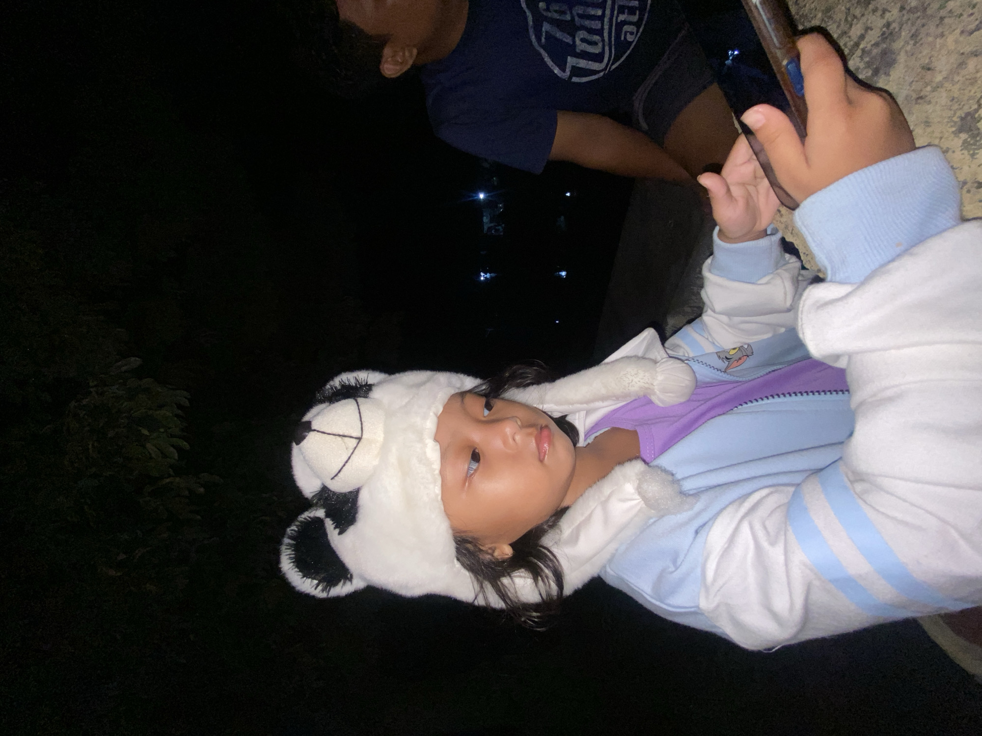 A young child is seen wearing a panda-ear hat and a light blue and white jacket. The child is holding a mobile phone in their hands. A second person is partially visible in the background, wearing a dark blue shirt. The photograph appears to have been taken at night, with the location possibly in Kalitengah, Indonesia, based on the prompt. The background is dark with some lights visible in the distance.