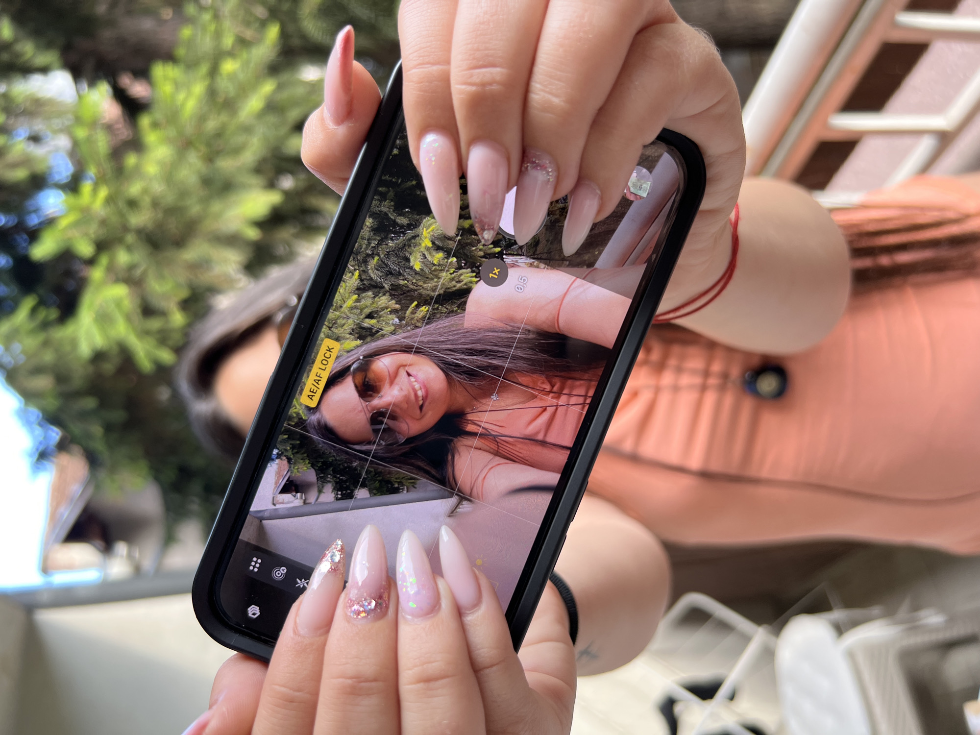 A woman with long, light brown hair and sunglasses smiles as she takes a selfie on her phone. She is wearing a peach-colored dress and has long, light pink acrylic nails with glitter accents. The phone screen displays the camera interface, showing a grid and zoom controls. In the background, blurred green foliage suggests an outdoor setting. The image has a shallow depth of field, with the woman and her phone in focus and the surroundings out of focus. The scene appears to be a balcony or outdoor area, likely in a residential setting.