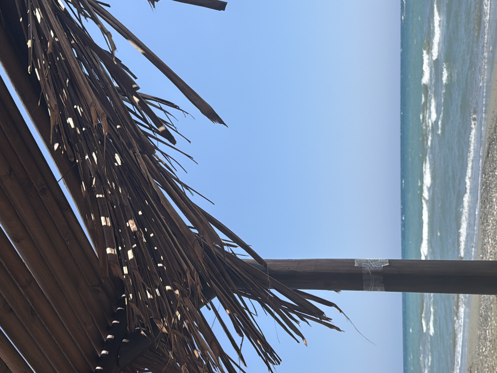 The image captures a section of a beach in Meneou, Cyprus, under a clear blue sky. In the foreground, a weathered wooden pole supports a thatched roof, likely part of a beach umbrella or shelter. The roof is made of dried palm fronds, with gaps allowing sunlight to filter through, creating dappled patterns. The fronds are frayed and show signs of age and exposure to the elements. A section of the wooden pole has been wrapped with clear tape. Beyond the shelter, the turquoise waters of the Mediterranean Sea stretch towards the horizon, with gentle waves breaking on the shore. The beach itself appears to be a mix of sand and small pebbles. No people are visible in the frame.