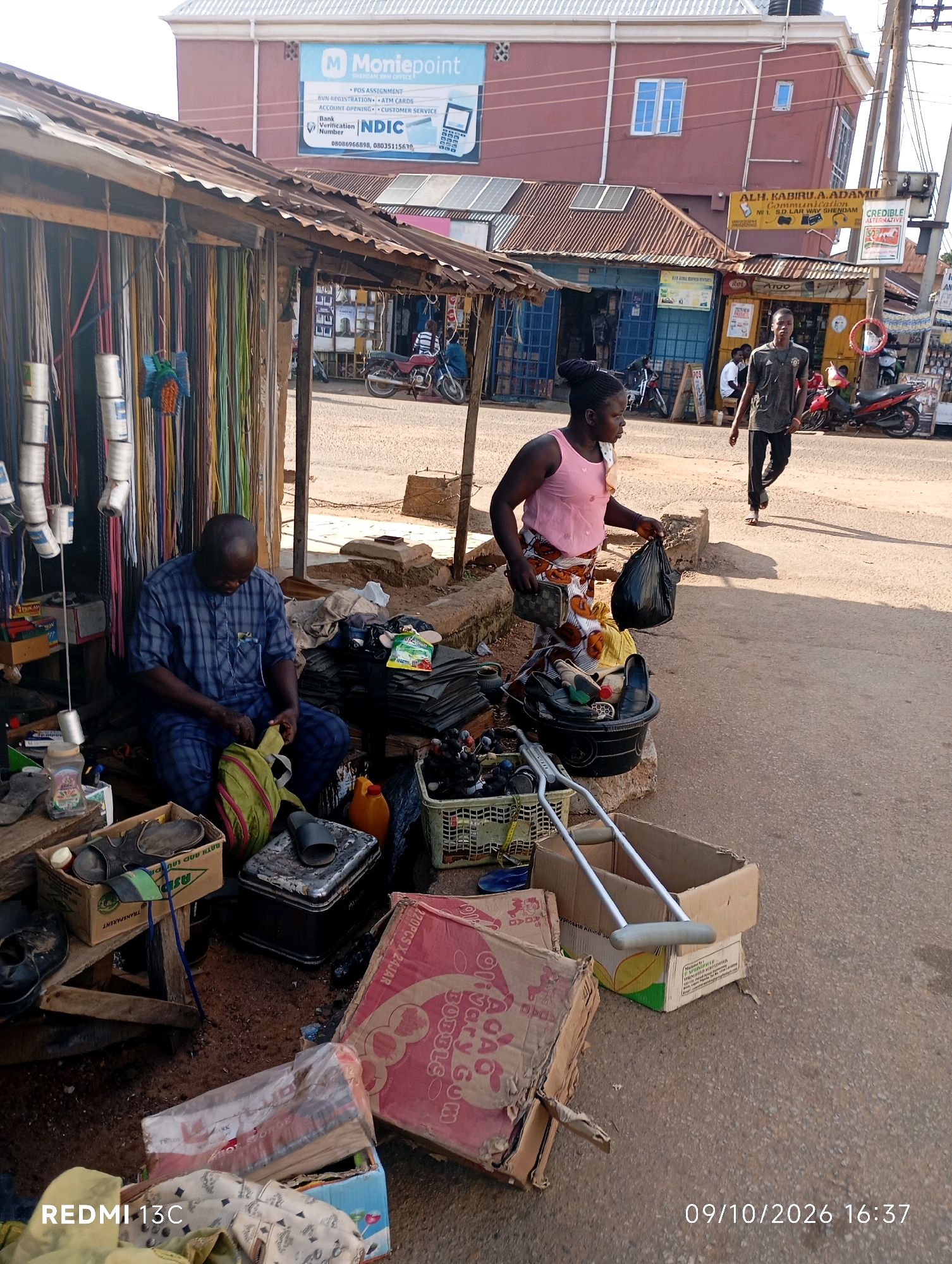 The image captures a street scene in Shendam, Nigeria, likely in front of a small shop or market stall. In the foreground, a man in a blue checkered shirt and pants is seated, appearing to be sorting through items in a green bag. To his right, a woman in a pink tank top and a patterned skirt is holding a black plastic bag and a small clutch purse. Various items are scattered around them, including shoes, boxes, plastic containers, and what appear to be industrial materials like electrical cords or ropes hanging from the stall's roof. A pair of crutches lies across a cardboard box in the mid-ground. In the background, two men are walking on the street, and several motorcycles are parked. A building with "Moniepoint" signage is visible further in the background, alongside other storefronts. The ground is paved with asphalt.