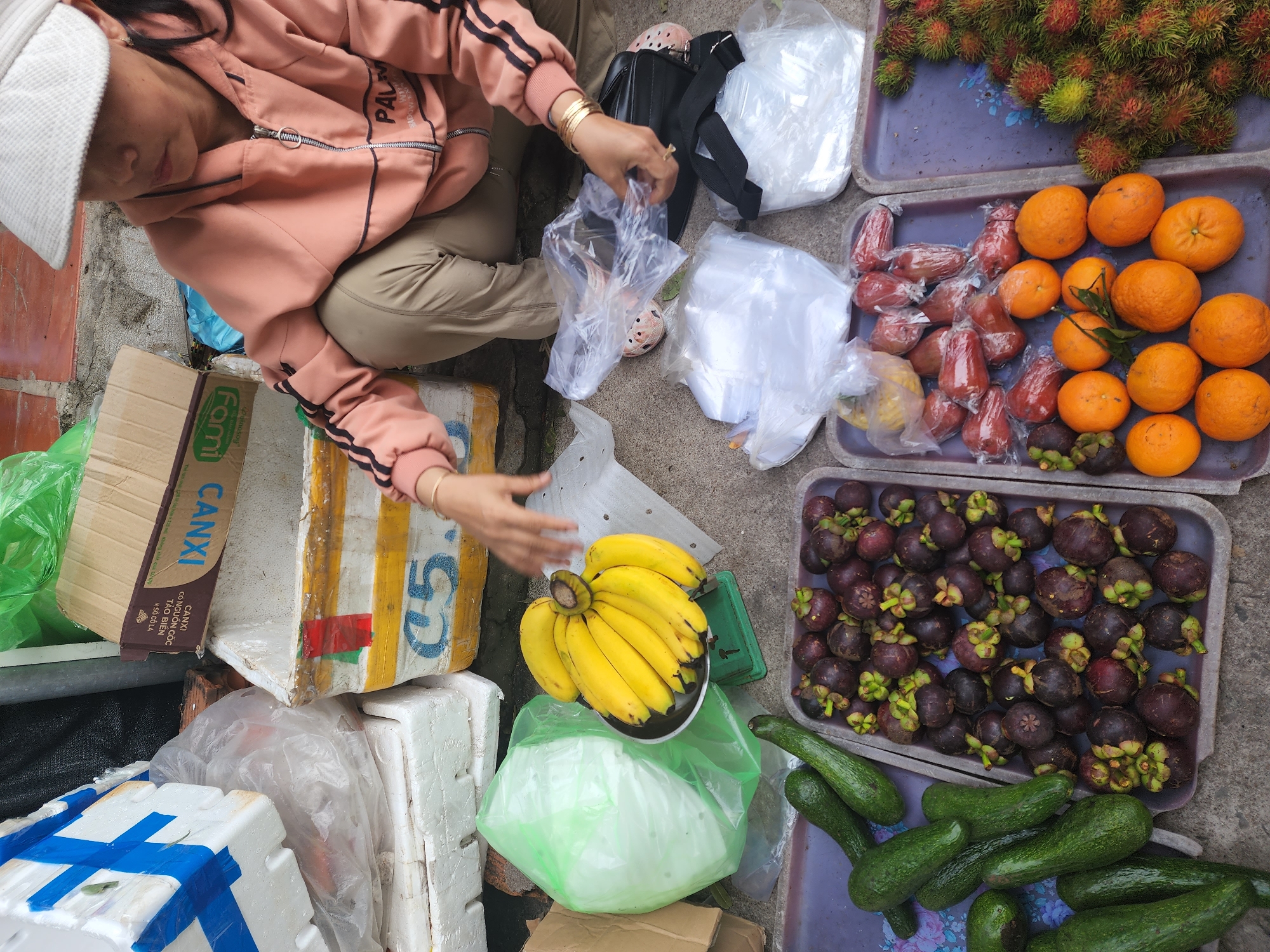 An outdoor market stall is depicted in Phu Thuy Ward, Vietnam. A woman wearing a peach-colored hooded jacket and tan pants is crouched down. She wears a white hat and is holding plastic bags. Various fruits are displayed on trays. In the foreground, a tray holds several avocados. To the right of the avocados is a tray filled with mangosteens. Further to the right, a tray contains oranges, guavas, and other small red fruits wrapped in plastic. A tray on the far right is overflowing with rambutans. In the background, a bunch of bananas is displayed on a small green scale. Several styrofoam boxes are stacked nearby, one with blue and white tape. A brown cardboard box is also visible. The ground is a rough, gray concrete surface.