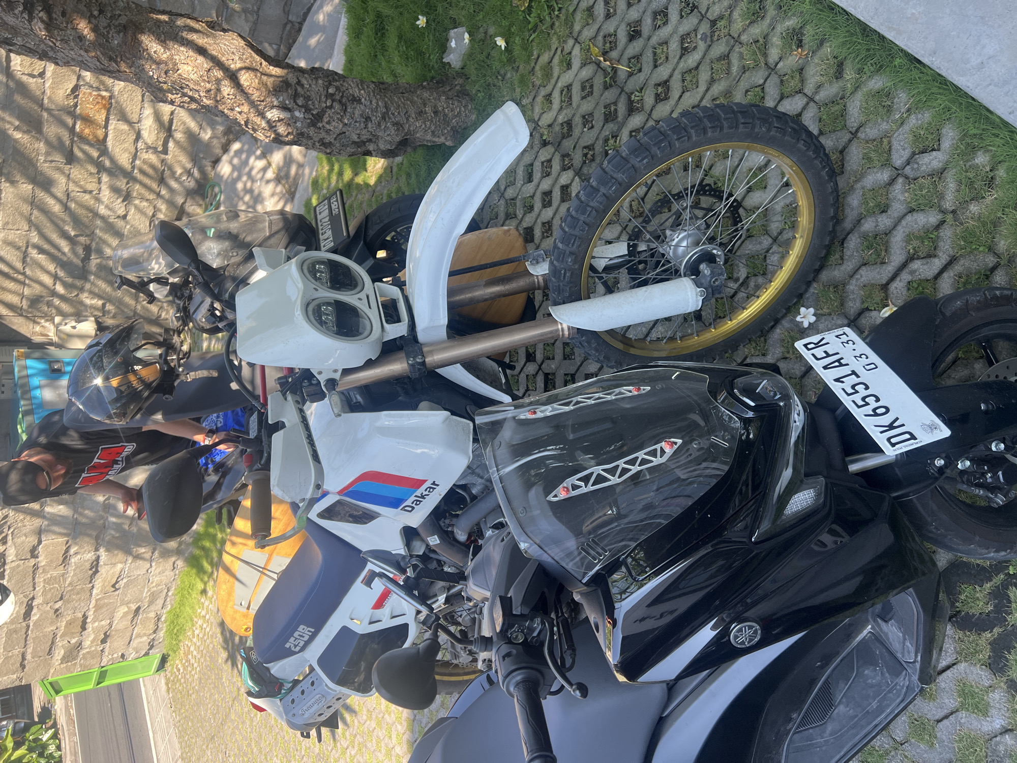 This image captures an outdoor scene in North Kuta, Indonesia, featuring two motorcycles parked on a paved area interspersed with grass, under bright sunlight. The lighting casts distinct shadows from a nearby tree, indicating a sunny day, likely morning or afternoon.
In the foreground on the right, a black Yamaha scooter, possibly a Yamaha NMAX, is parked. Its front panel prominently displays the Yamaha logo. A white Indonesian number plate, "DK 6551 AFR" with the registration/expiration date "03.24" (March 2024), is affixed to its rear.
Behind the black scooter, a white and blue adventure motorcycle is parked. It features a distinctive "Dakar" decal on its side panel and "250R" on its seat, suggesting an adventure touring bike, possibly a modified BMW GS F 650 Dakar or a similar style. This motorcycle is equipped with dual round LED headlights, a large white front fender, and a gold-rimmed front wheel with knobby off-road tires. A yellow surfboard is strapped horizontally to the rear of this bike. Its number plate, "DK 5827 FH" with "03.23" (March 2023), is partially visible. The "DK" prefix on both number plates confirms the location as Bali, Indonesia.
A person is partially visible on the far left, standing near the white motorcycle. They are wearing a black t-shirt with red lettering (appears to be "RIP") and a black cap or helmet. Another helmet rests on the handlebars of the black scooter. The background includes a stone wall, the textured trunk of a tree, and small white frangipani flowers scattered on the grass.