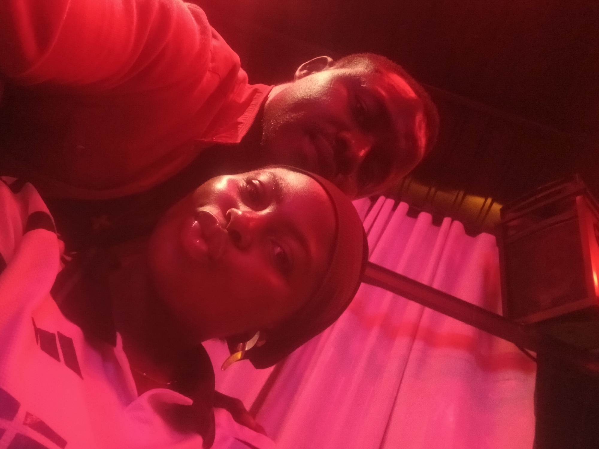 The image shows two people, likely a couple, in a dimly lit environment with a strong red or pink light cast. The woman in the foreground is wearing a head wrap and earrings and is looking directly at the camera. The man is behind her and also looking at the camera. Behind the couple is a curtain, a metal bar, and what appears to be a light fixture.
Based on the location prompt Rukuba Road, Nigeria, it is likely this image was taken in or around a business or residential area within the city of Rukuba Road, Nigeria. Due to the red or pink lighting, it is likely they are inside a building at night.