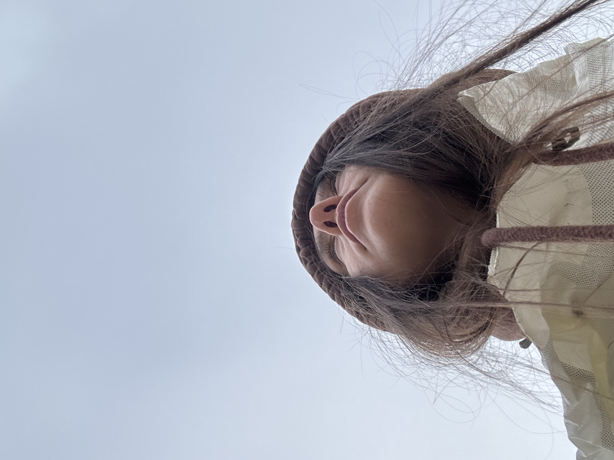 A low-angle perspective captures a person wearing a brown hooded garment and a light-colored jacket against a light blue or gray sky. The person's face, visible from the nose and upwards, is framed by brown hair with some stray strands. The location appears to be outdoors, implied by the sky as the backdrop. The context of Zelenogradsk, Russia, is not directly evidenced within the visual information.