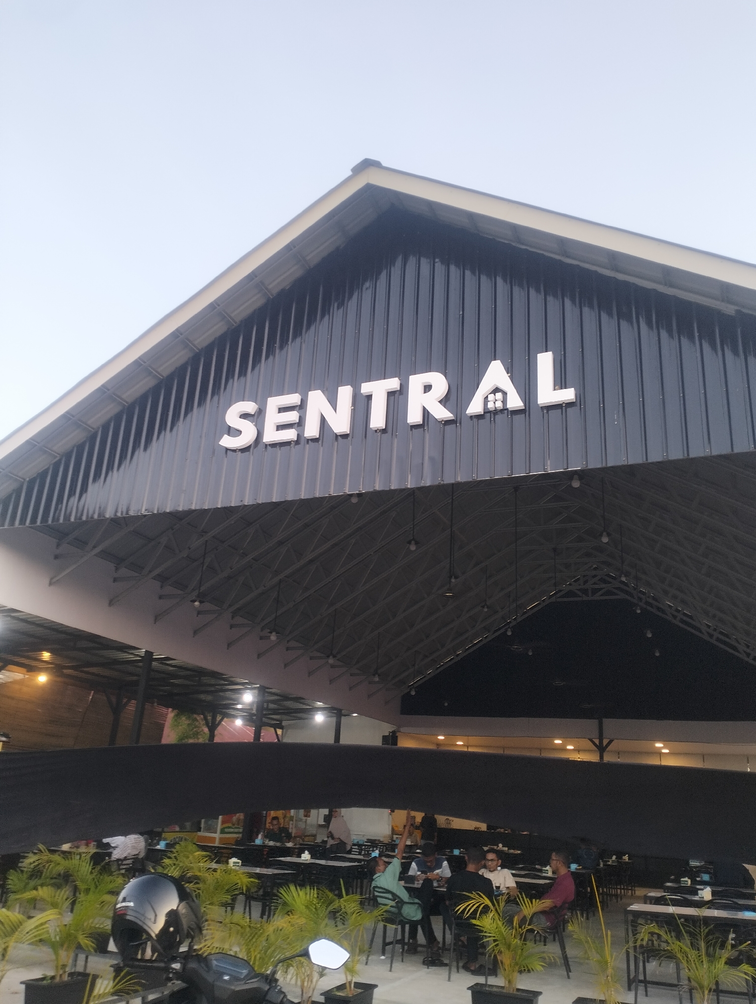 The image captures the exterior and a partial interior view of a restaurant with a prominent "SENTRAL" sign mounted on a dark, corrugated metal facade. The sign's letters are large and white, with the 'A' stylized to resemble a house with a roof and chimney. Below the sign, an open-air dining area is visible, supported by a metal truss structure. Several tables and chairs are set up, with some patrons seated and others standing. Small potted plants with green fronds are strategically placed around the seating area. A black motorcycle helmet sits on a stand in the foreground, reflecting the overhead lights. The restaurant appears to be located in Lhokseumawe, Indonesia, as indicated by the surrounding context. The overall scene suggests a casual dining establishment, possibly an outdoor eatery or a restaurant with an open-air section.