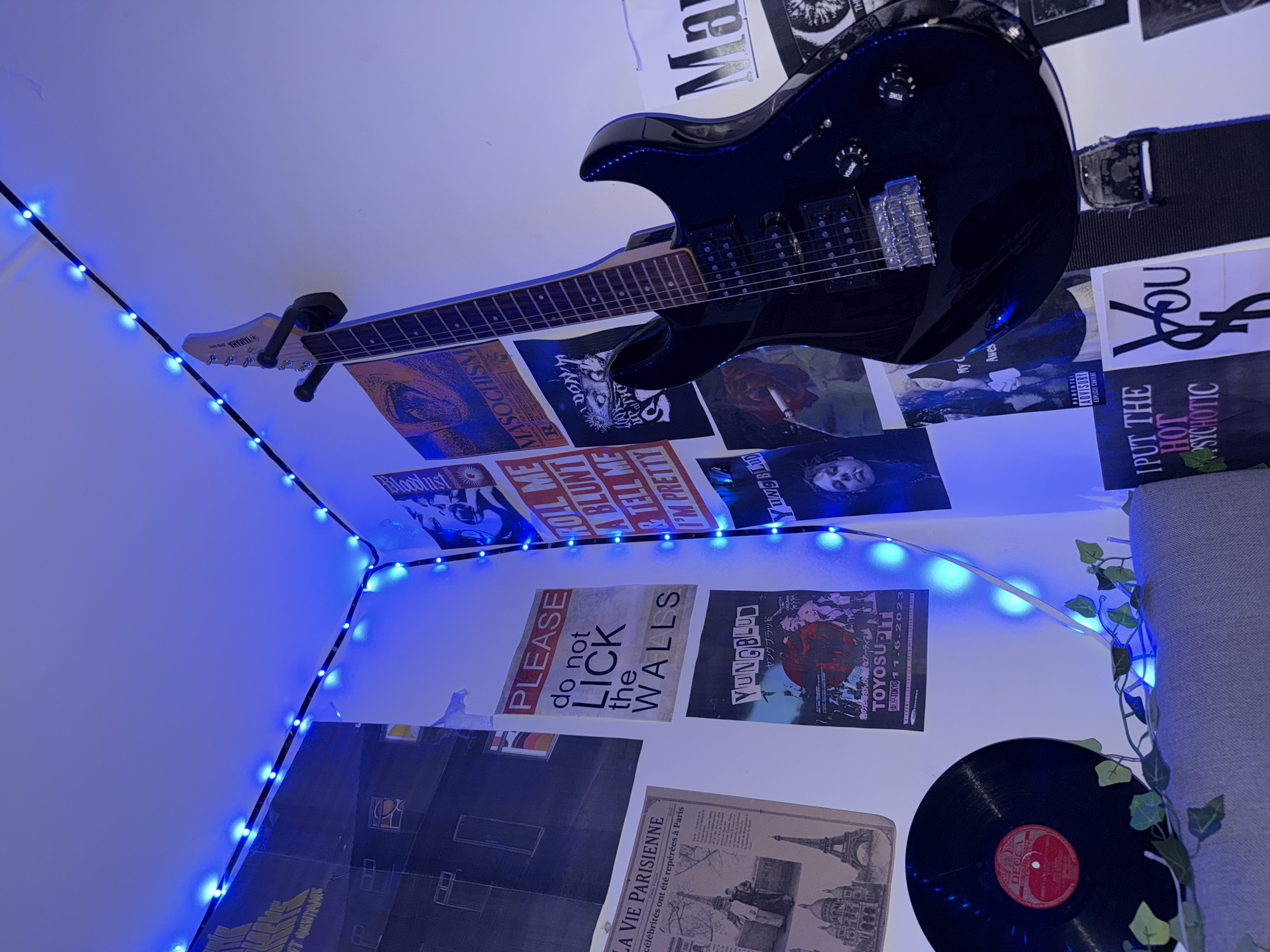 A black electric guitar hangs on a wall adorned with various posters. Blue LED string lights outline the corner of the room. On a grey surface below the guitar, a black vinyl record with a red label is visible, along with a decorative vine. The posters display a range of imagery and text, including what appears to be album art, band posters, and graphic designs. One poster, a prominent red and white one, reads "PLEASE do not LICK the WALLS". Another poster features the text "YUNGBLUD". The environment appears to be a bedroom or a dedicated space for music enthusiasts. There are no people visible in the image. The location is not specified as Oslo, Norway.