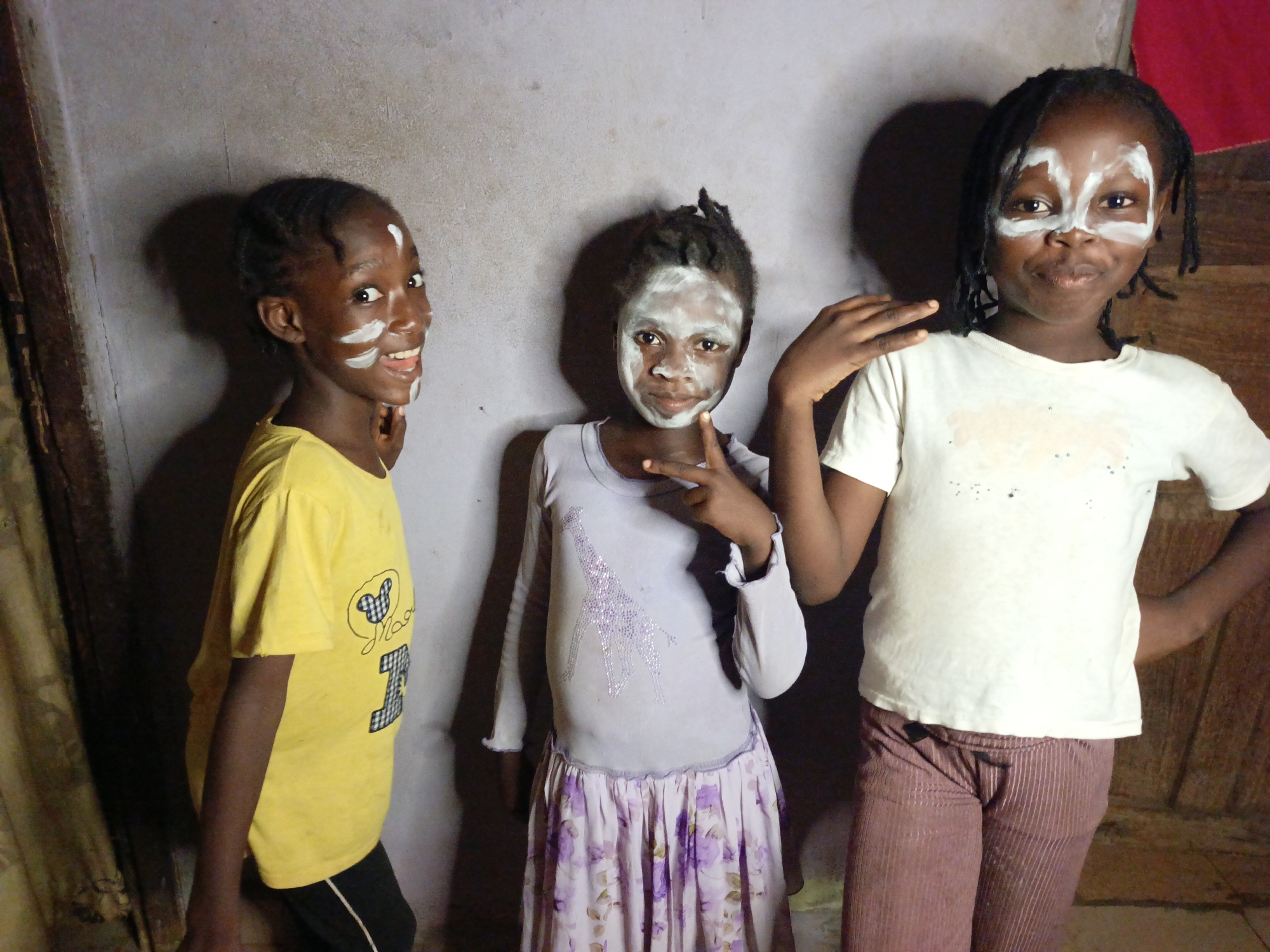 Three young children, likely girls, of African descent, are pictured indoors, posing against a light grey, textured wall. All three have white paint or makeup applied to their faces in distinct patterns, suggesting a playful activity or cultural celebration.
The child on the left wears a yellow short-sleeved t-shirt with a partially visible design featuring a Mickey Mouse head outline and the number "13". She smiles broadly, looking towards the camera, with white paint accentuating her forehead, cheeks, and chin.
In the center, a child wears a light purple long-sleeved shirt adorned with a sparkling giraffe design and a light purple floral skirt. Her face is more extensively covered in white paint, and she holds up two fingers in a "peace" or "V" gesture.
The child on the right is dressed in a white short-sleeved t-shirt and maroon, vertically striped pants. White paint frames her eyes in a mask-like design. She offers a subtle smile, with one hand resting on the shoulder of the middle child and the other on her hip.
The children appear joyful and engaged with the camera. The background includes the plain wall and a dark wooden door or panel on the far right. Prominent shadows on the wall suggest indoor lighting.