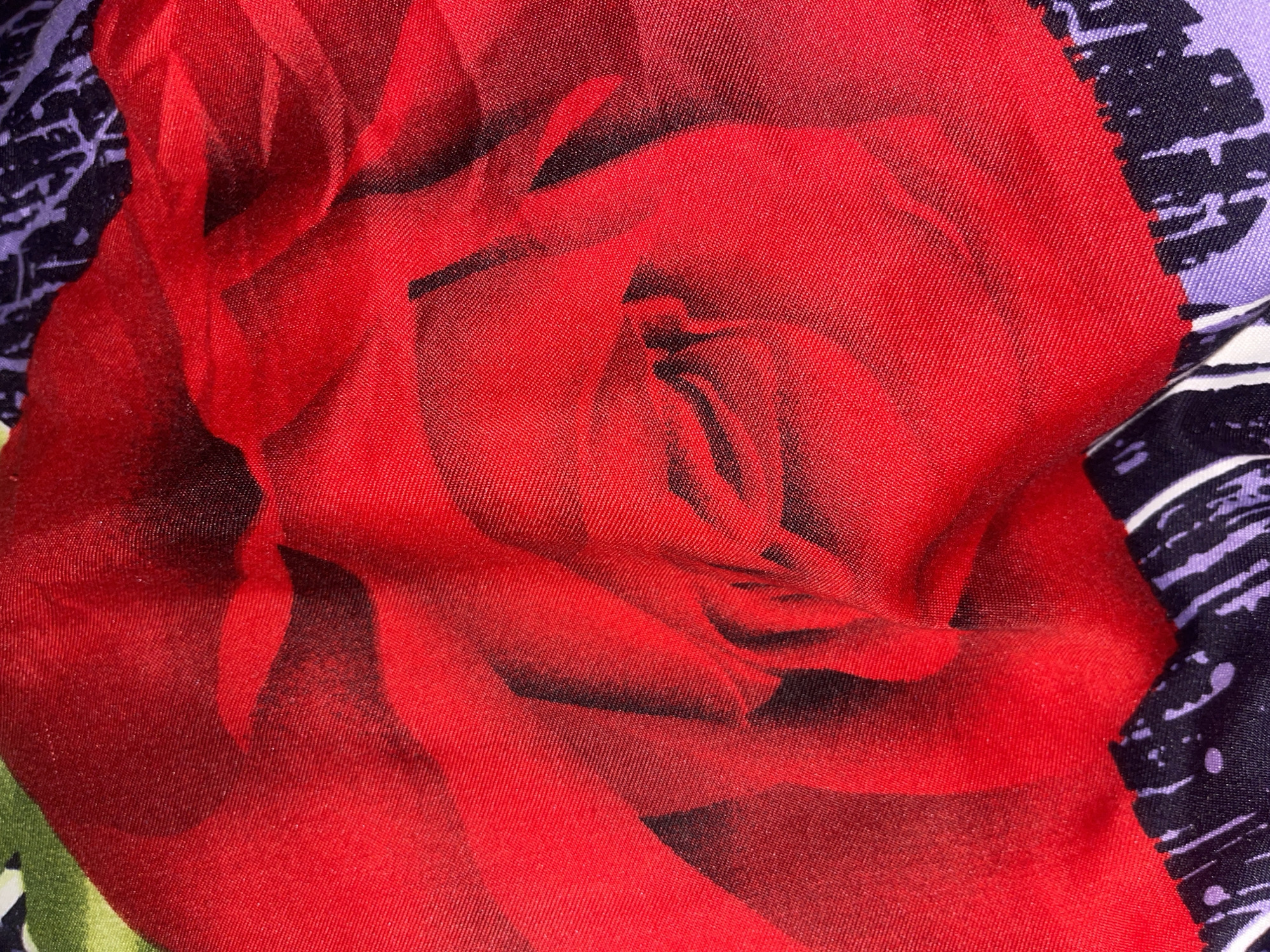 The image is a close-up, high-angle shot of a red rose pattern on fabric. The rose is depicted in various shades of red, with darker areas suggesting shadows and depth within the petals. The fabric appears to be satin or a similar material with a visible weave texture. In the lower portion of the image, the pattern transitions to a border with abstract black and purple designs. The lighting is somewhat harsh, creating highlights and deep shadows on the fabric's surface, which is gently creased and folded. There are no visible people or elements of the Minsk, Belarus environment present in this image.