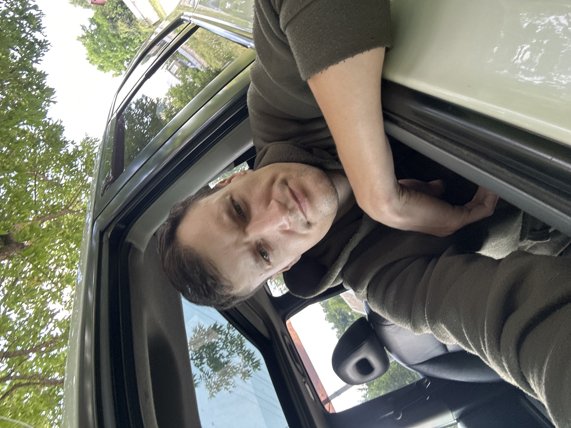 A man is taking a selfie from inside a car, leaning out of the open window. He is wearing a dark olive green hooded sweatshirt. The interior of the car is visible, including a black leather seat and headrest. Sunlight streams through the sunroof, and green foliage from trees is visible outside the car. The image appears to have been taken during the daytime.