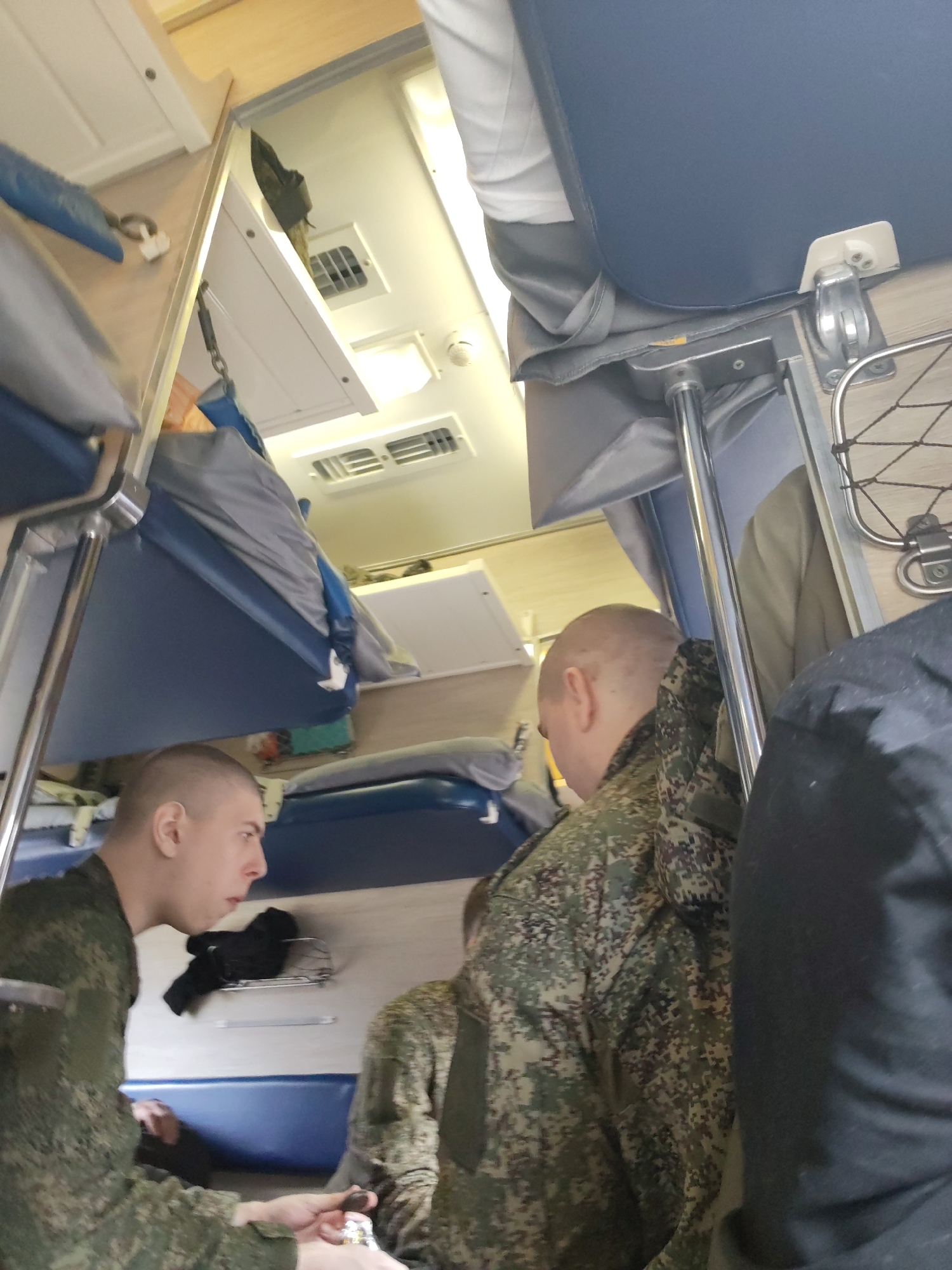 The image captures the interior of a crowded train carriage, consistent with a Russian Platskart class sleeper car. Two young men with shaved heads are prominent subjects, both wearing pixelated green and brown camouflage uniforms, suggesting military personnel. One man is seated on a lower bunk on the left, looking downwards and holding an object in his hands. The other uniformed man is seated opposite him, seen from the back and side. A third person, whose back is partially visible on the far right, wears a black jacket.
The setting is characterized by rows of bunk beds with blue upholstery and grey bedding, supported by metal frames. The interior features white walls, light-colored wooden surfaces, overhead lighting, and ventilation grilles. Small metal storage racks are affixed to the walls, one holding a black item. A dark backpack is visible on an upper shelf. The individuals appear to be engaged in train travel, with neutral expressions. The military uniforms and the style of the train car are strong indicators of a Russian context, aligning with the provided location of Shushkovo, Russia. No discernible text is visible.