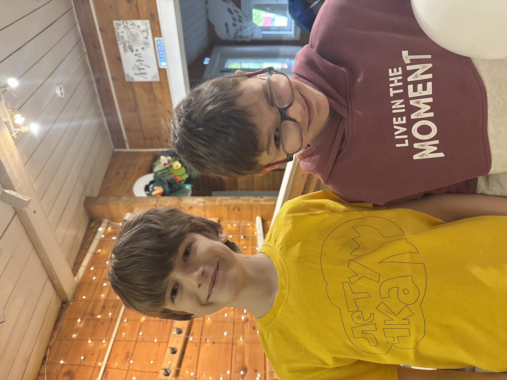 Two boys are posing indoors with festive lighting. The boy in the yellow t-shirt is smiling with shoulder-length brown hair and is positioned in the foreground. Behind him is a boy wearing glasses and a burgundy hoodie with the words "LIVE IN THE MOMENT". Behind them, the interior features wooden walls decorated with string lights, a wall sign in Cyrillic, and a decorative arrangement of a plush figure. It is not possible to determine the location of the city of Moscow, Russia.