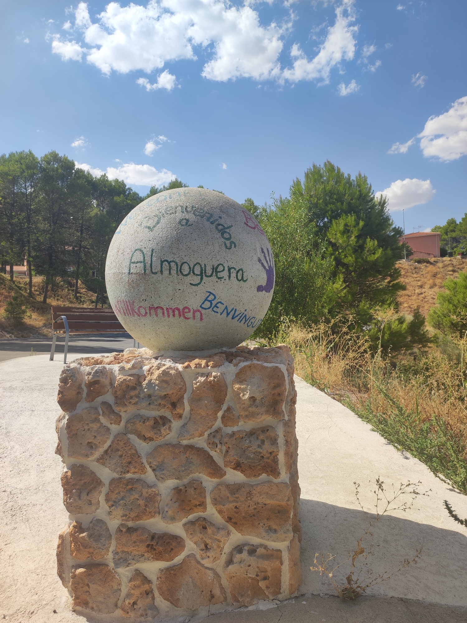 This is an eye-level, medium shot of a concrete sphere decorated with painted text and a handprint, set on a stone pedestal. The sphere is located outdoors on a sunny day, with a clear blue sky and scattered white clouds. Green trees and a hillside with dry vegetation are visible in the background. A wooden bench with a metal frame is situated to the left of the sphere. The text on the sphere appears to be a welcome message in multiple languages, including "Bienvenidos," "Almoguera," "Willkommen," and "BENVINGUDA." A purple handprint is painted on the upper right portion of the sphere. The pedestal is constructed from rough-hewn, light-brown stones. The ground is paved with a light-colored concrete surface, and dry, sparse vegetation grows along the edge. A building with a red roof is visible on the hill in the background. The image appears to be taken in Almoguera, Spain, as indicated by the text on the sphere.